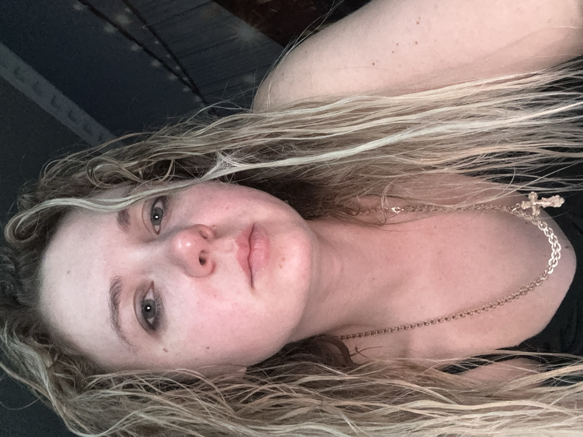 A close-up, slightly low-angle, indoor selfie shows a young white woman with long, wet, blonde hair that frames her face. She is looking directly at the camera with a neutral expression. Her eyes are open, and her mouth is closed. She has light skin with some redness, and faint dark circles under her eyes. She is wearing a black top and a gold necklace with a cross pendant. The background is dark and out of focus, with some horizontal lines visible, suggesting a ceiling or structure. The lighting is dim, with a warm cast. The woman's face and upper body occupy most of the frame.