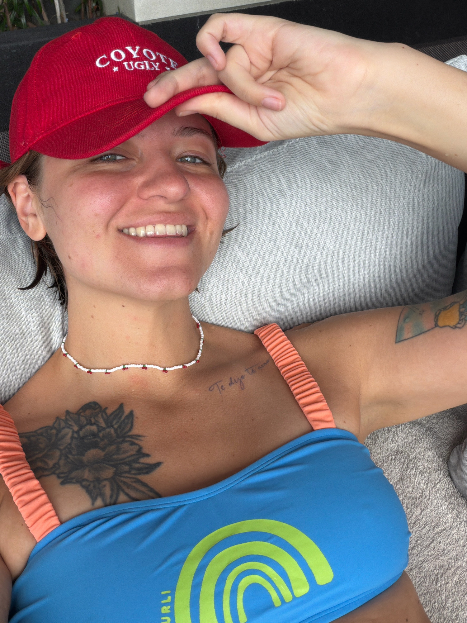 A close-up, over-the-shoulder selfie shows a woman lying down and smiling. She is wearing a red baseball cap with "COYOTE UGLY" embroidered in white letters. She has a beaded necklace with red and white beads and green accents around her neck. Her right arm is resting on a gray textured surface, possibly a cushion or sofa, and her hand is holding the brim of her cap. She is wearing a blue bikini top with orange straps, and a faint tattoo of flowers is visible on her chest. Another tattoo is visible on her upper right arm. The background appears to be a lounging area with a gray textured surface. The image is captured in a bright, outdoor setting, likely during the daytime. This scene is consistent with the relaxed atmosphere found in resorts or private villas in Hua Hin City Municipality, Thailand, where such leisurely activities are common.