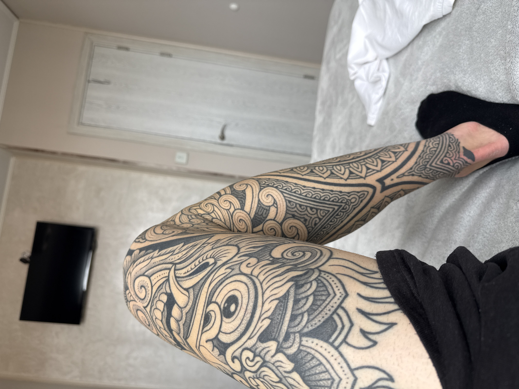 The image depicts a person's leg and arm covered in intricate black tattoos resting on a grey bedspread. The leg is bent at the knee. A black sock covers the foot. A white piece of fabric is on the bed. The upper portion of the arm is partially obscured by black clothing. In the background, a wall features a closed white door. A black television is mounted on the wall. The setting appears to be an interior space, possibly a hotel room. There is no verifiable information to suggest this image was taken in Sofia, Bulgaria.