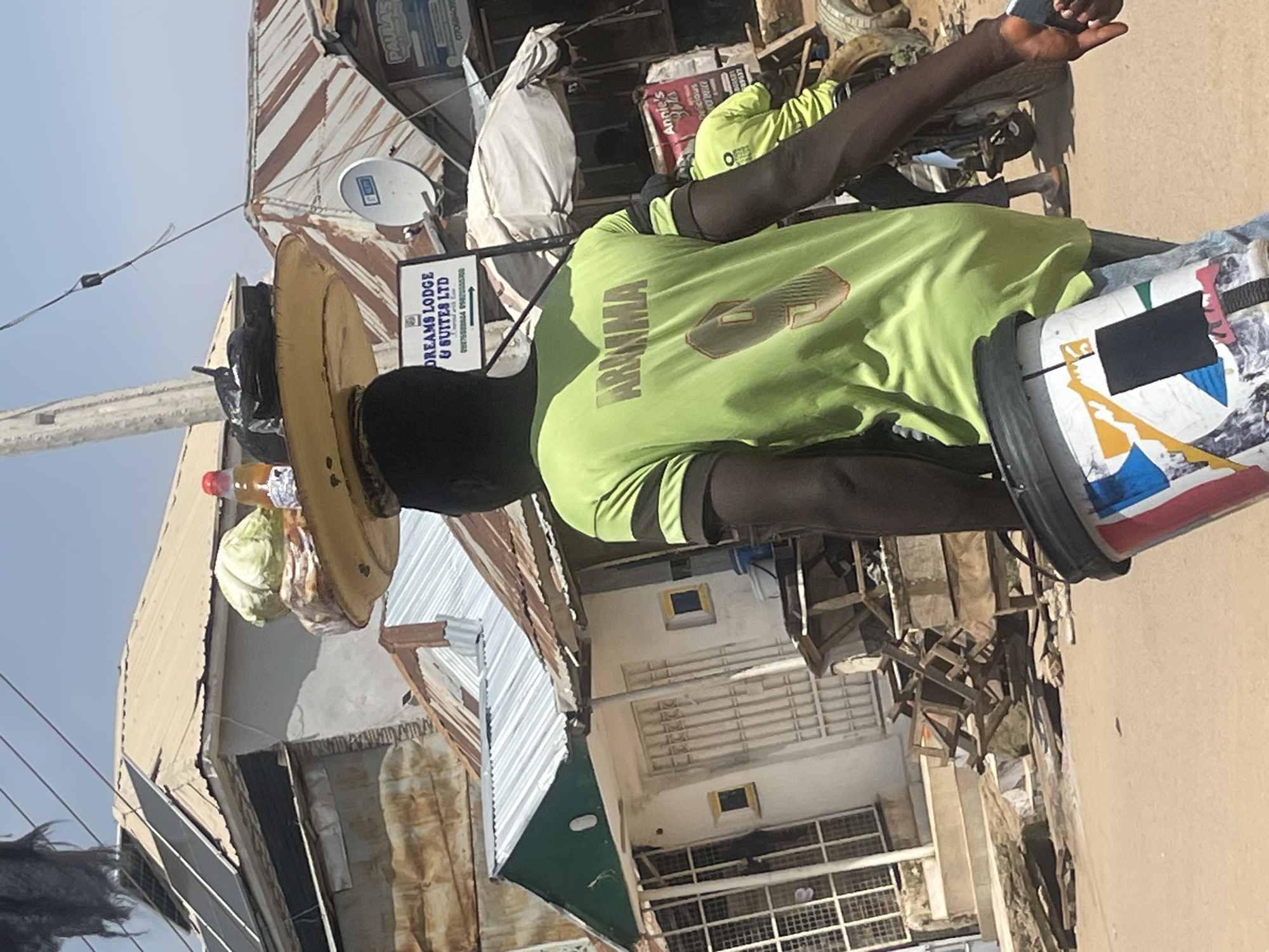 The image depicts a street scene in Abuja, Nigeria, likely on a sunny day given the shadows and bright lighting. In the foreground, a young man is walking away from the camera, carrying a large, colorful bucket in his right hand and what appears to be a mobile phone in his left hand. He is wearing a bright green t-shirt with the word "ARUMMA" and the number "9" printed on the back, with darker green stripes on the sleeves.
Balanced on the man's head is a large, flat, yellow tray laden with items. These include a head of cabbage, a bottle of liquid with a red cap, and a black plastic bag. The tray itself is round and appears to be made of metal or plastic.
In the background, the environment suggests a developing urban area. Buildings with corrugated metal roofs are visible, some appearing weathered and in disrepair. One building has solar panels installed on its roof. A sign reading "DREAMS LODGE & SUITES LTD" is prominently displayed. A motorcycle is partially visible to the right, with another person wearing a similar green t-shirt on it. The ground is a paved or dirt road. The overall impression is of a bustling, everyday scene in a Nigerian city.