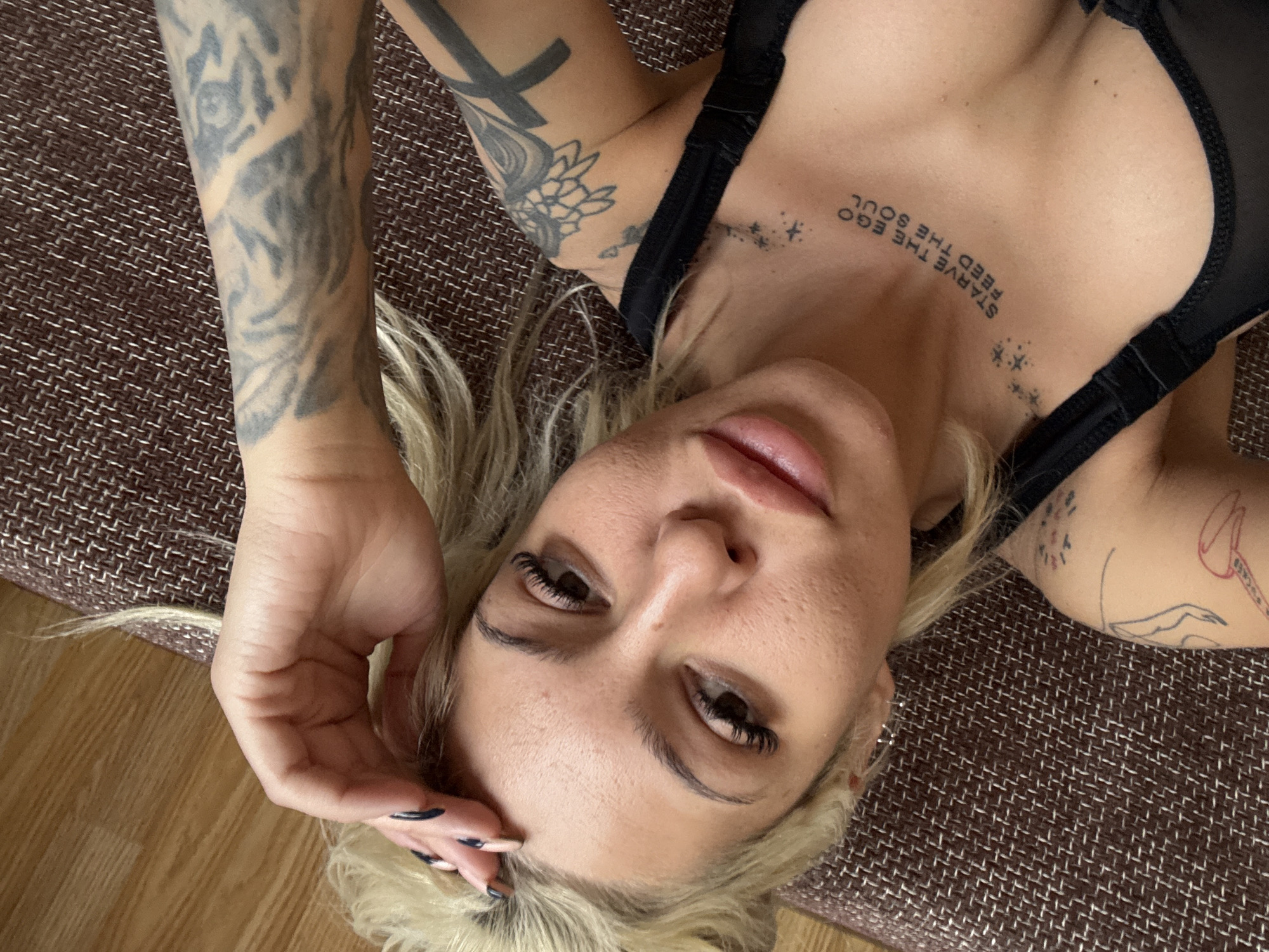 A woman with blonde hair and tattoos is lying down on a textured surface. She is wearing a black top. One arm is raised, and her hand is near her forehead. The background includes a wooden floor. The location is Unknown, Unknown.