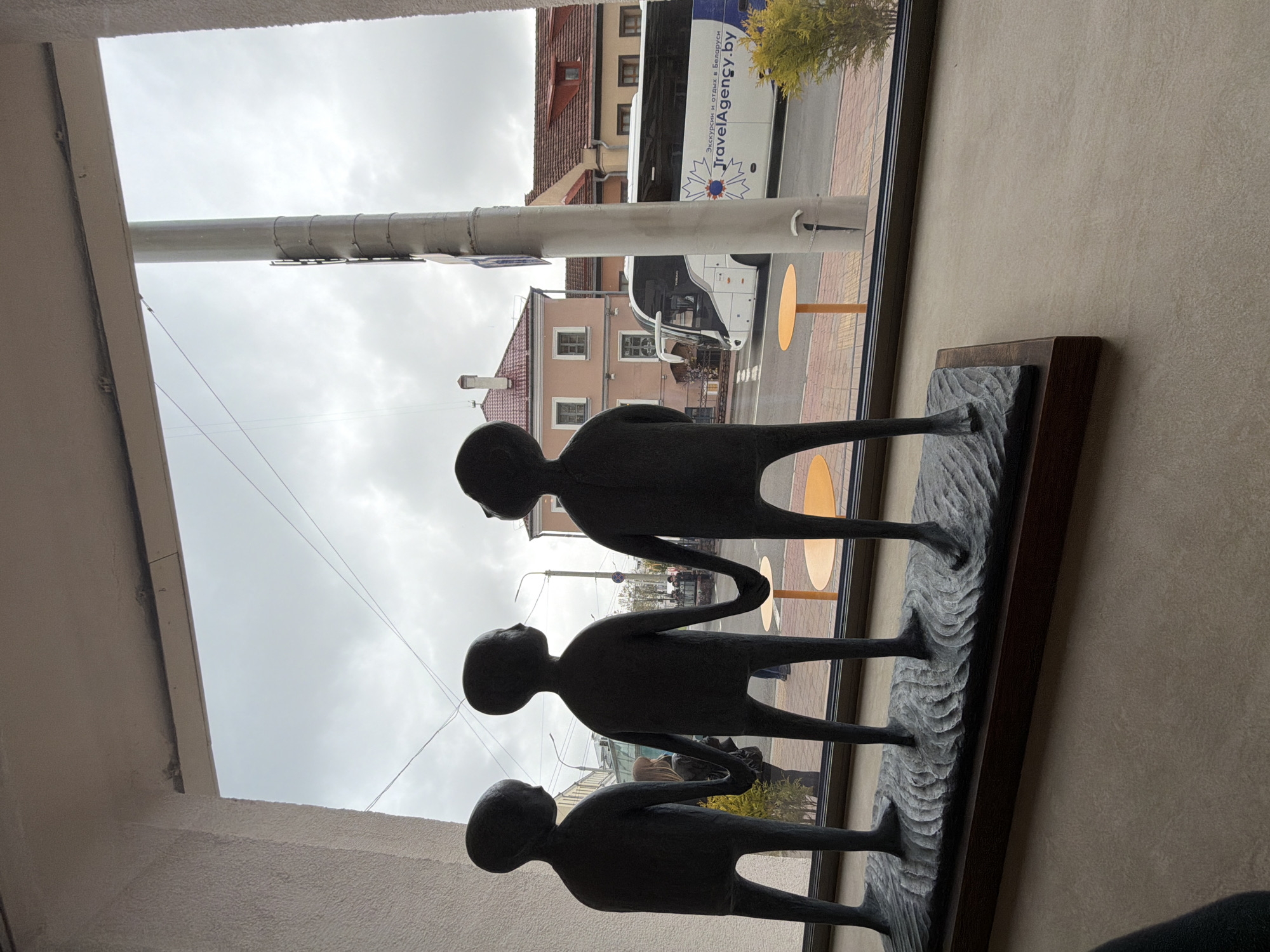 A low-angle, interior shot depicts a sculpture of three stylized human figures in a row, holding hands, with a city view through a nearby window. The sculpture is dark and mounted on a dark wooden base with a rough, carved texture beneath the figures' feet. Through the window, buildings with red-tiled roofs are visible on an overcast day. A bus is also visible, bearing the inscription "TravelAgency.by" and featuring an advertisement in an unknown language. The bus and buildings suggest the scene is in the city of Unknown, Unknown. In front of the buildings, orange circular objects are visible. The photo is taken from indoors, with a white wall or frame visible at the bottom and left of the shot.