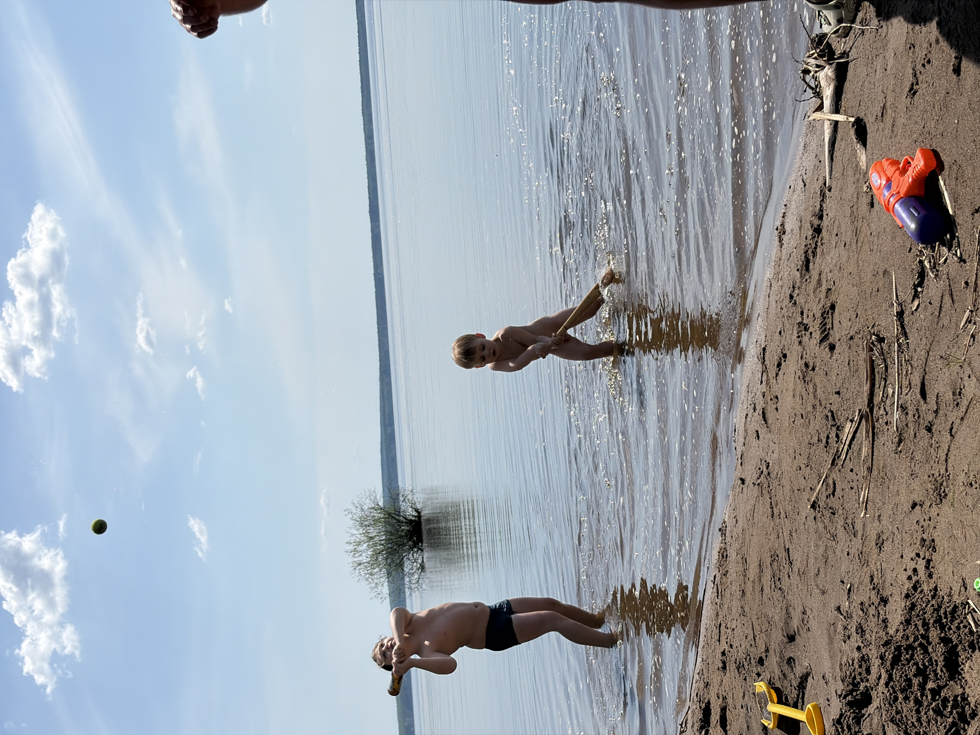 The image depicts a waterfront scene. A boy wearing dark-colored swim trunks stands in shallow water at the edge of the shore, holding an object to his mouth. A younger, naked boy is also in the water, further out from the shore. The water has visible ripples and reflections. On the sandy shore, there are objects including a bright orange and blue toy, a yellow toy shovel, and bits of debris like twigs and grass. The sky is blue with scattered white clouds. The perspective is rotated 90 degrees counter-clockwise. The location is Zabegaevo, Russia. A tennis ball is in the sky. A bush is visible in the water.