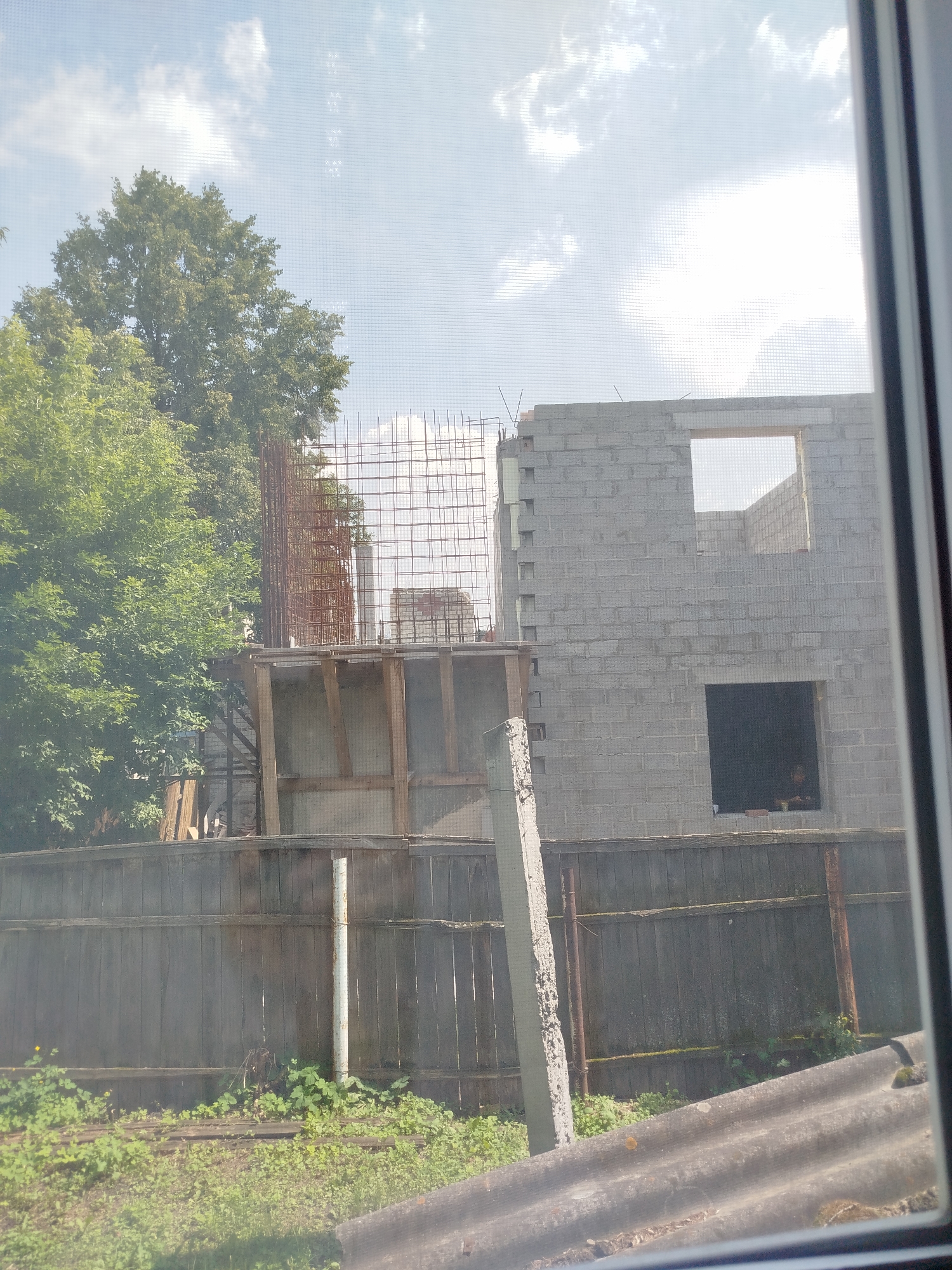 The image depicts a construction site viewed through a window screen. The foreground shows a weathered wooden fence with some overgrown greenery and a portion of a corrugated roof. Behind the fence, there is a partially constructed building made of grey concrete blocks, with openings for windows and doors. Reinforcement bars are visible, indicating ongoing construction. To the left of the building, a large green tree stands. The sky is visible with clouds and a bright sun. The location is identified as Zavodoukovsk, Russia.
