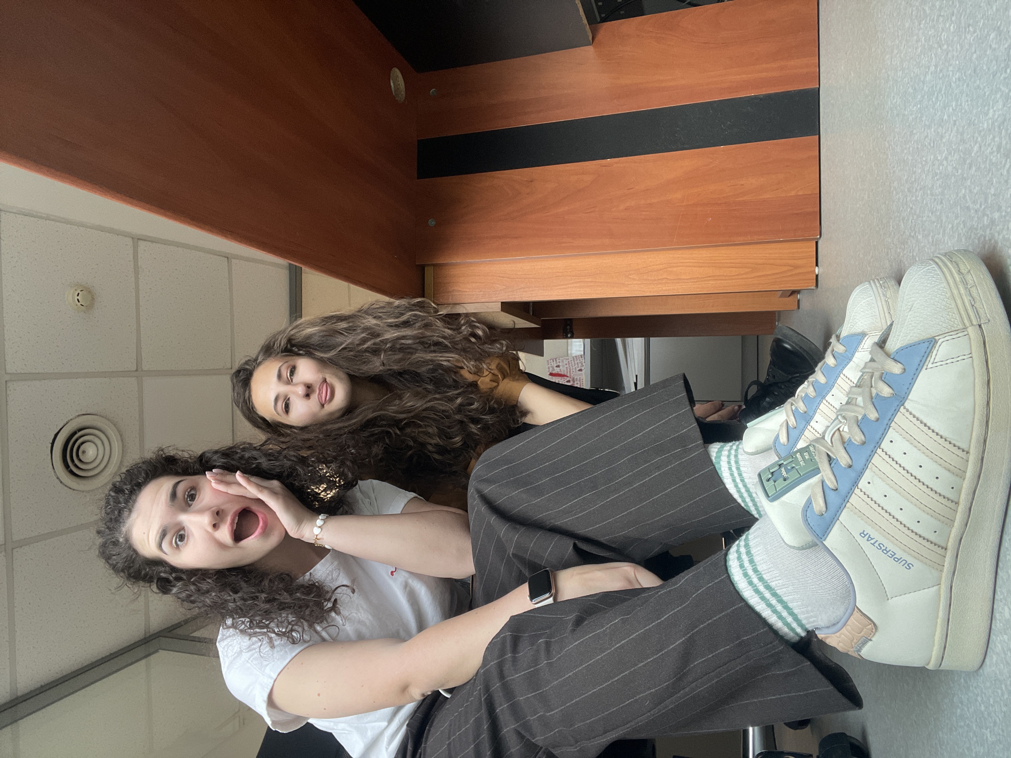 Two young women are in an office environment. One wears a white t-shirt and pinstripe trousers and gestures with her hand near her open mouth, as if calling out, or feigning surprise. The other woman behind her has long, wavy brown hair and is sticking her tongue out, suggesting a playful mood. They appear to be seated under an office desk made of brown wood. The woman in pinstripes has white sneakers with blue stripes and green-and-white striped socks. It is not possible to determine if the location is in Sofia, Bulgaria from the image.