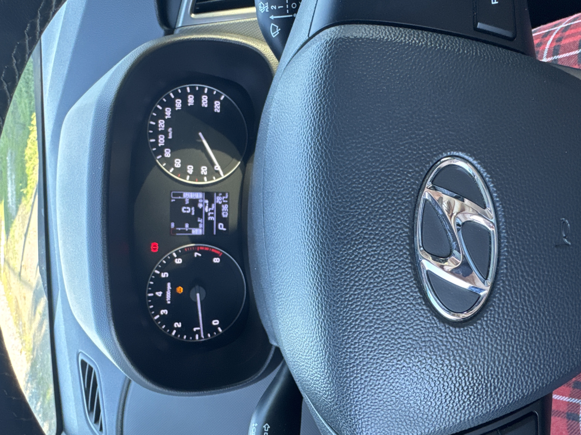 The image shows the interior of a Hyundai car, focusing on the steering wheel and dashboard. The dashboard features a digital display with information such as speed (0 km/h), gear (P), and trip distance (10361 km). The tachometer shows 0 rpm, and there's an orange illuminated warning light, likely for the engine or tire pressure. The steering wheel has the Hyundai logo prominently displayed in the center. To the left of the steering wheel, a turn signal lever is visible, and to the right, a windshield wiper control stalk is present. The background outside the car is blurred but suggests a sunny outdoor environment with some greenery. The image is likely taken by someone in the driver's seat. The car is parked in Starokhalilovo, Russia.