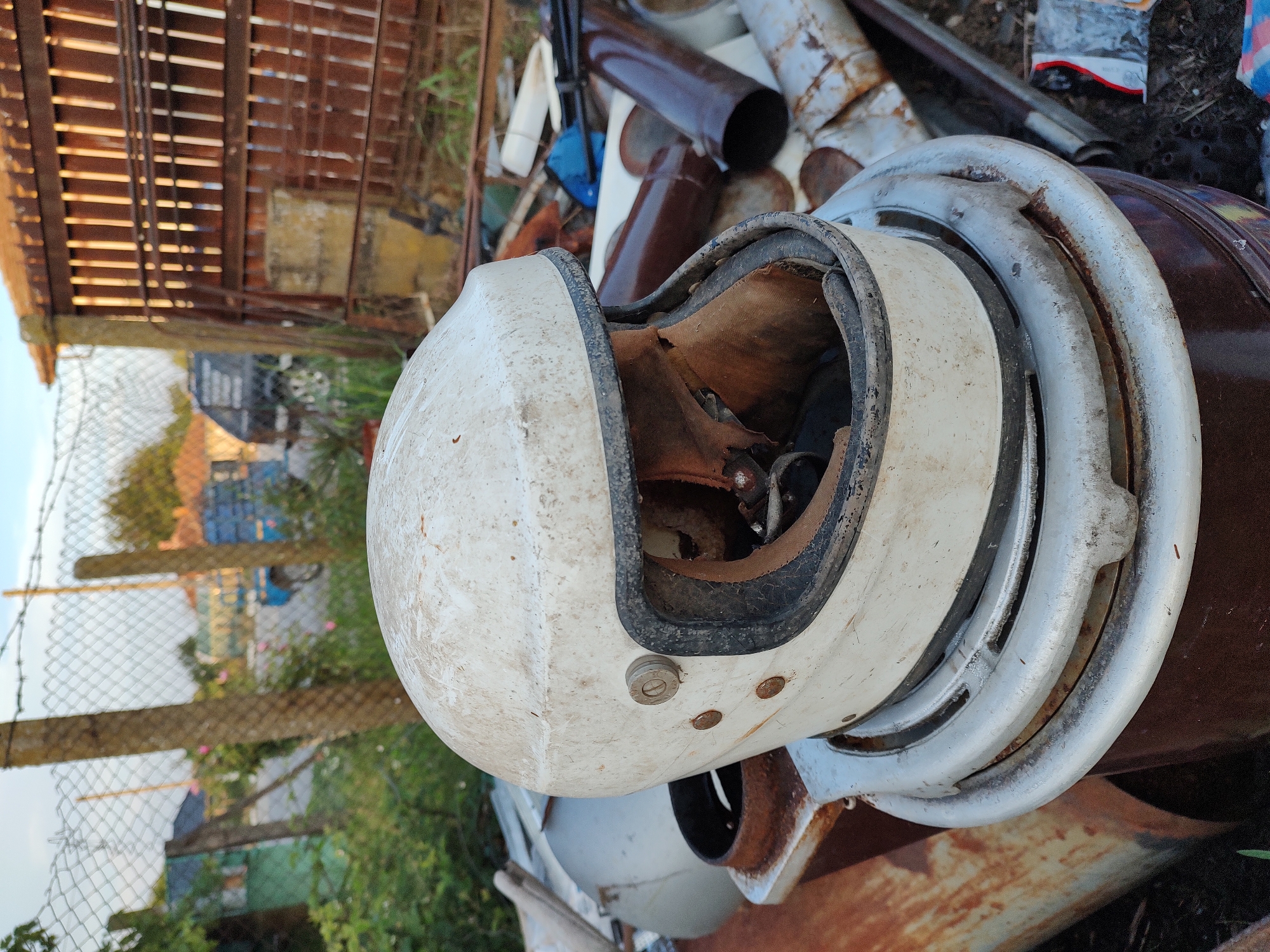A weathered, white motorcycle helmet sits atop a rusty metal barrel in an outdoor area. The helmet is dirty and the interior lining is torn. The background shows a chain-link fence, wooden posts, overgrown vegetation, and a rustic wooden structure. In the distance, buildings, a blue truck, and a dark vehicle are visible. The scene appears to be in Babuk, Bulgaria, and suggests a junkyard or an abandoned outdoor storage area.