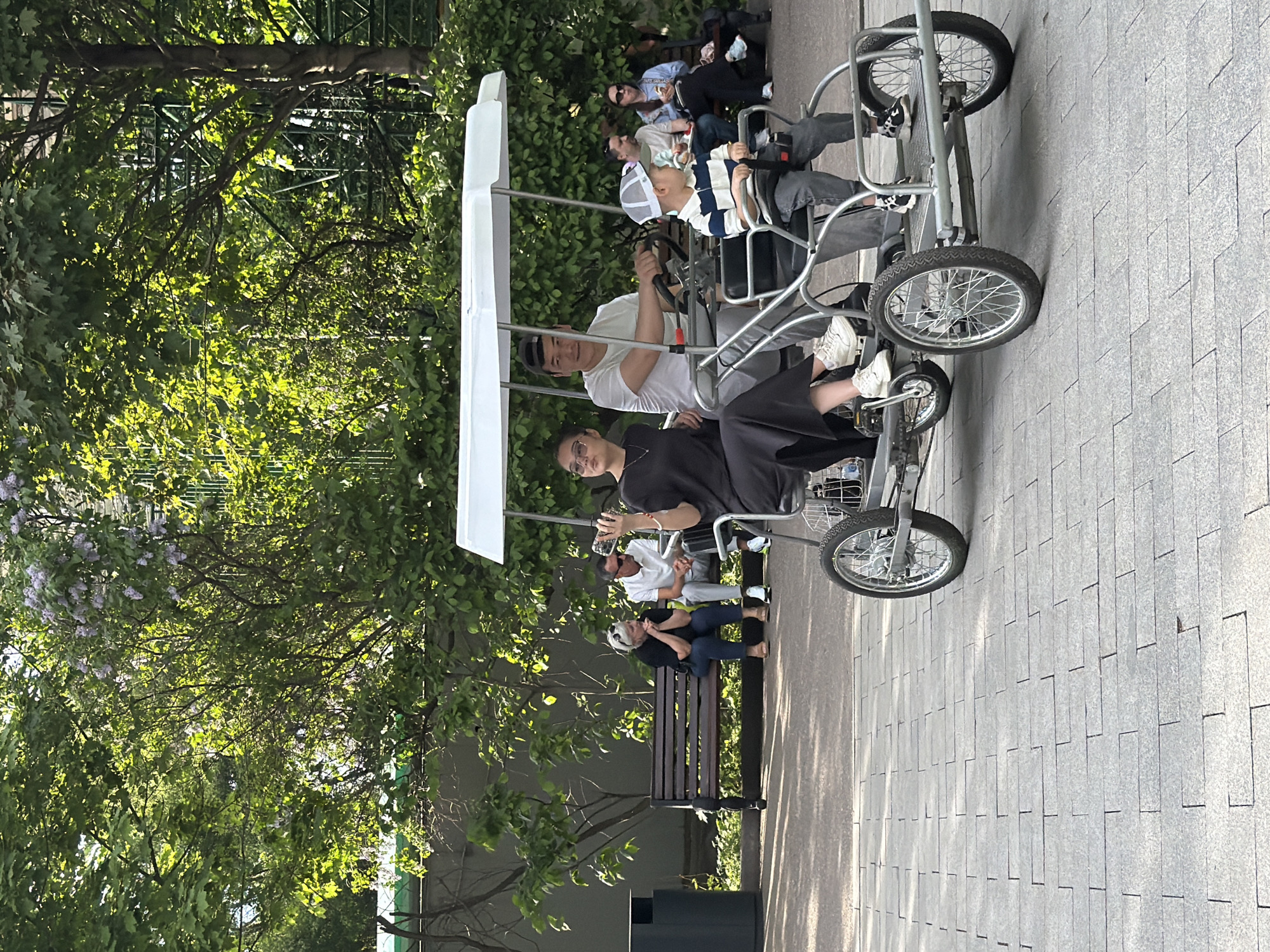 The image depicts a group of people in Moscow, Russia, riding a multi-person bicycle on a paved path in a park-like setting. The path appears to be constructed of rectangular paving stones. A white-roofed bicycle has four wheels and is occupied by four people, including adults and a child. Two individuals are seated in the front, each with their own set of pedals, while two other individuals are in the back. Behind and to the left of the bicycle are several people seated on park benches, under trees. The environment includes trees with green foliage, a trash can, and a possible low fence or structure in the background. The time of day appears to be midday, with bright sunlight casting shadows on the path.