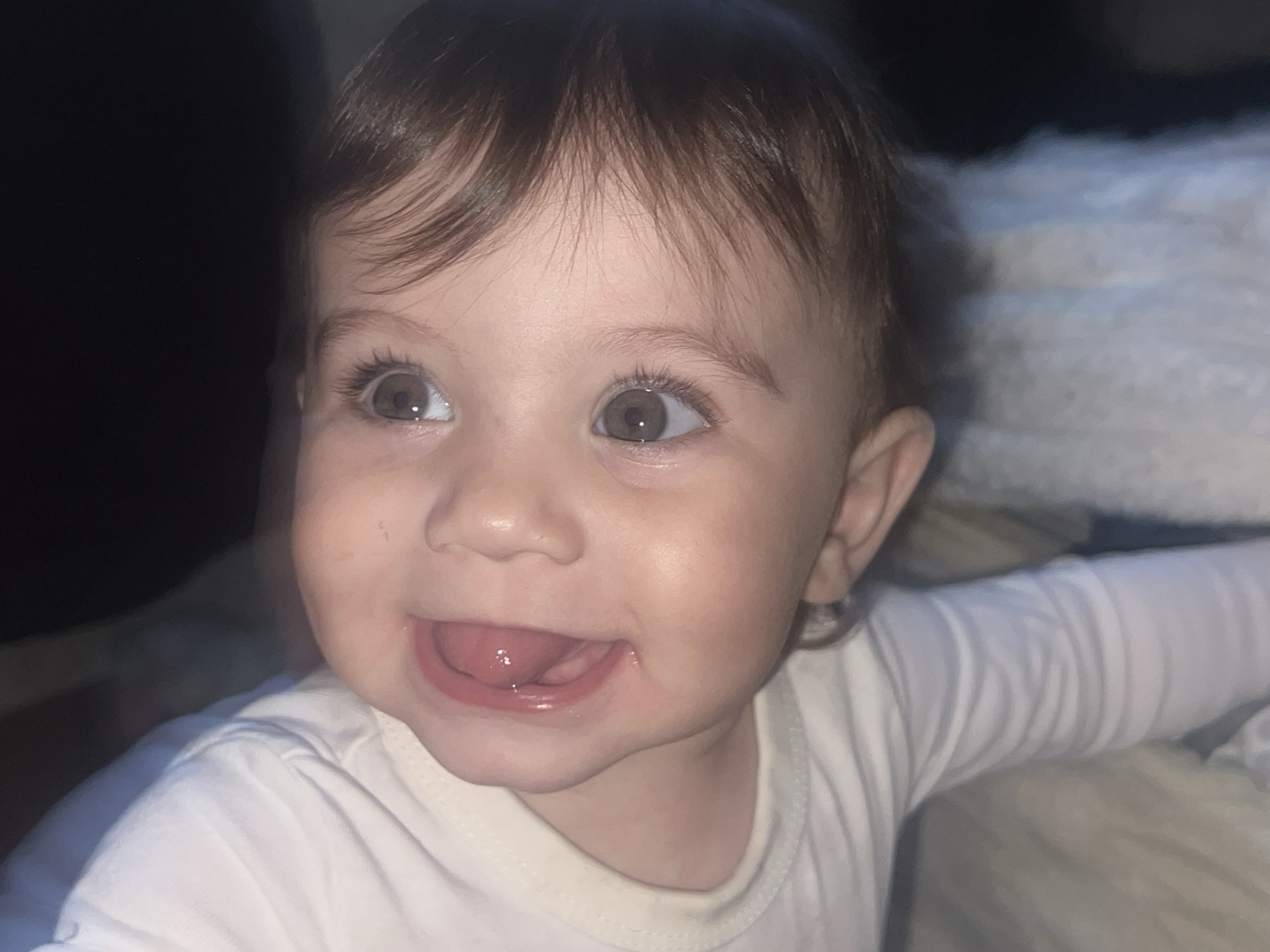 A close-up shot captures a baby with brown hair and large, gray eyes looking off to the left and slightly upward. The baby is wearing a white long-sleeved shirt. The background is partially dark, and a soft white blanket or sheet is partially visible. There is no visible information suggesting the location is Elena, Bulgaria.