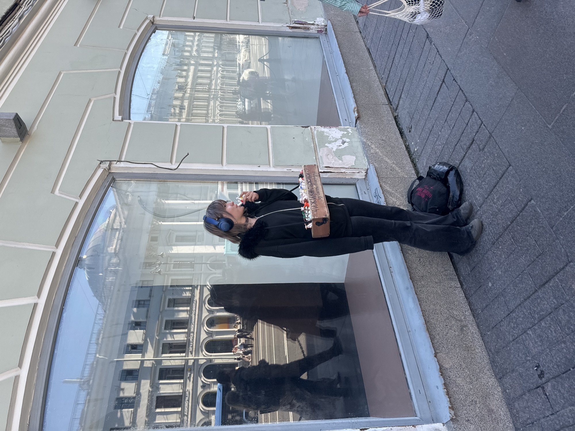 A young woman with headphones stands on a sidewalk in Saint Petersburg, Russia, holding a wooden box filled with trinkets and appearing to be eating. The box has Cyrillic writing on it, reading "Чижик-Пыжик" (Chizhik-Pyzhik), a reference to a famous St. Petersburg monument. She wears a black zip-up hoodie with a fur-lined hood, black pants, and sneakers. A black bag rests at her feet. The background features a building with large display windows reflecting a street scene with passing pedestrians and vehicles. The architecture suggests a historic urban environment.