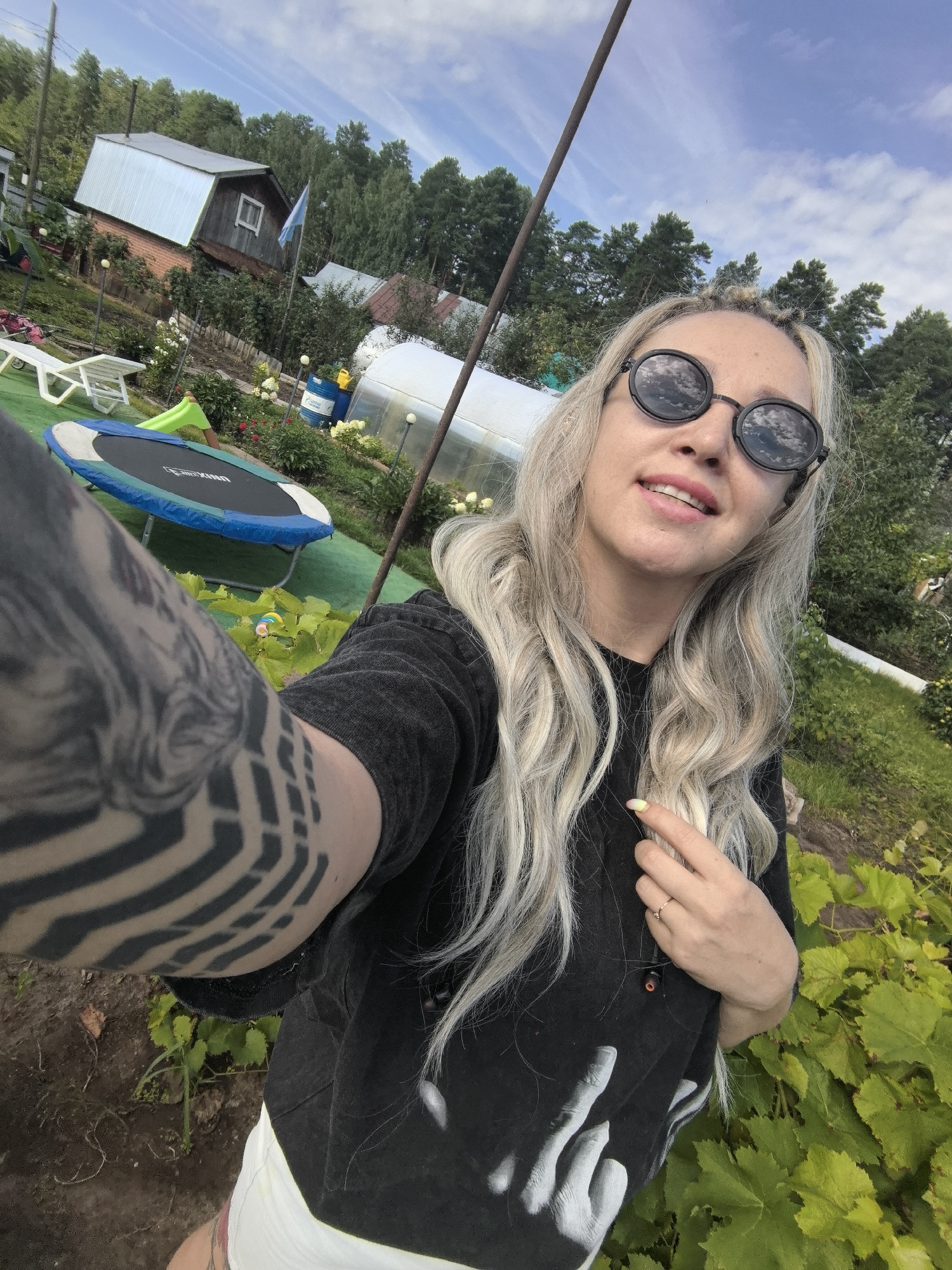 The image is a selfie taken outdoors, likely in a rural or suburban setting. A young woman with blonde hair and sunglasses is in the foreground, holding her phone to take the picture. She is wearing a dark t-shirt and white shorts. Behind her, a trampoline is visible, along with a greenhouse and a small building that appears to be a house. Trees and vegetation form the background. The sky is partly cloudy. The location is identified as Noviy, Russia.
