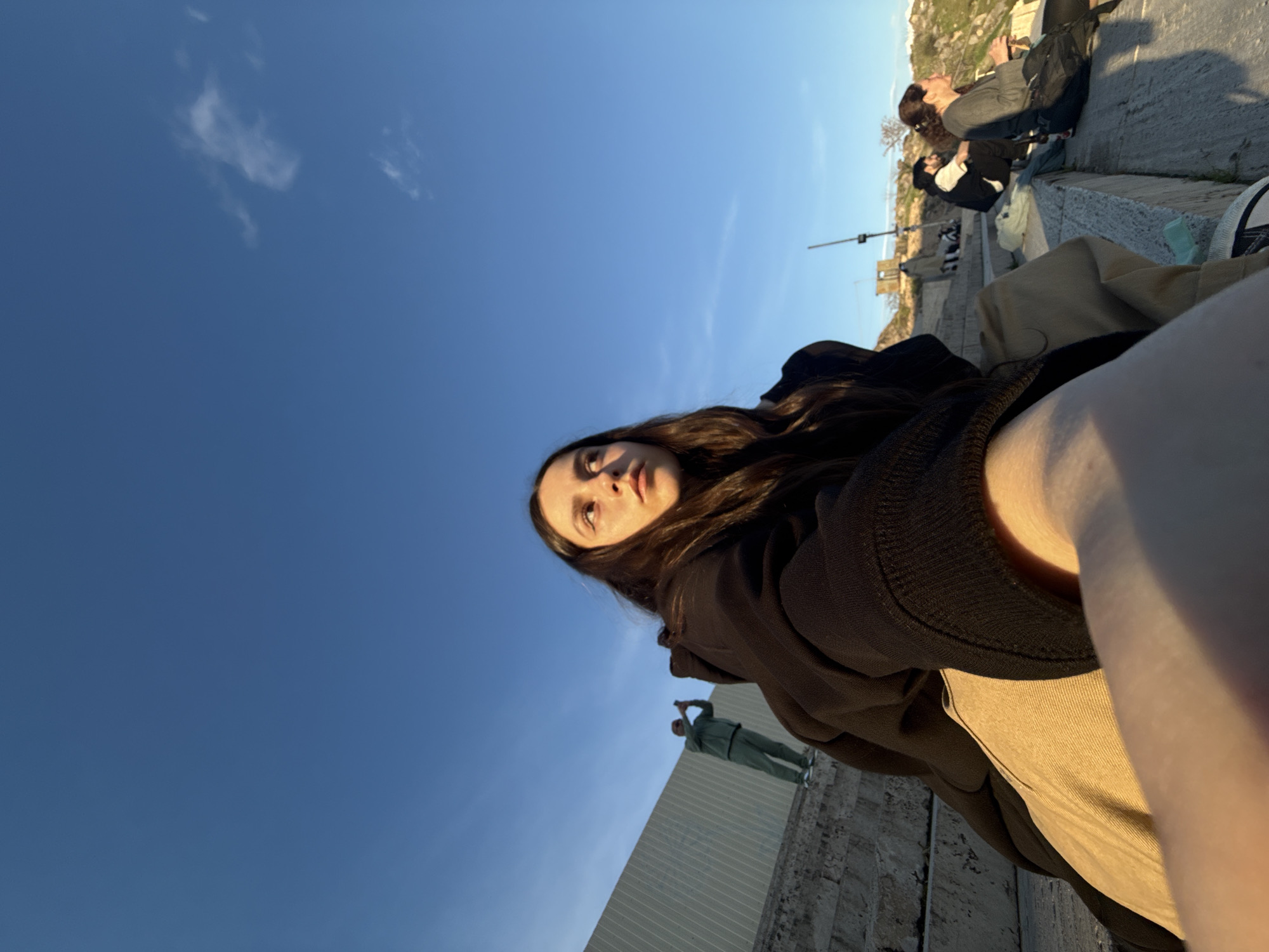 A low-angle selfie captures a young woman looking upwards into a vast, clear blue sky, bathed in the warm light of golden hour. She has long, dark, wavy hair and is wearing a dark, possibly black, long-sleeved top or hoodie, with a light beige or khaki garment visible on her lower body. Her face is well-lit, conveying a calm and contemplative expression.
The setting is an outdoor public space, likely a viewpoint or architectural site in Yerevan, Armenia. Concrete structures, ledges, and steps define the foreground and mid-ground. In the mid-distance, a person in a light green tracksuit stands on a wall, appearing to hold an object up. Further in the background, several other individuals are visible relaxing, seated on the concrete steps. One person wears a grey top and has a black backpack. The atmosphere suggests a leisurely afternoon or evening.
The vibrant blue sky dominates the upper portion of the frame, adorned with a few sparse, wispy white clouds, indicating clear and pleasant weather. The strong sunlight casts soft shadows and highlights on the subjects and environment. No discernible text is visible in the image.