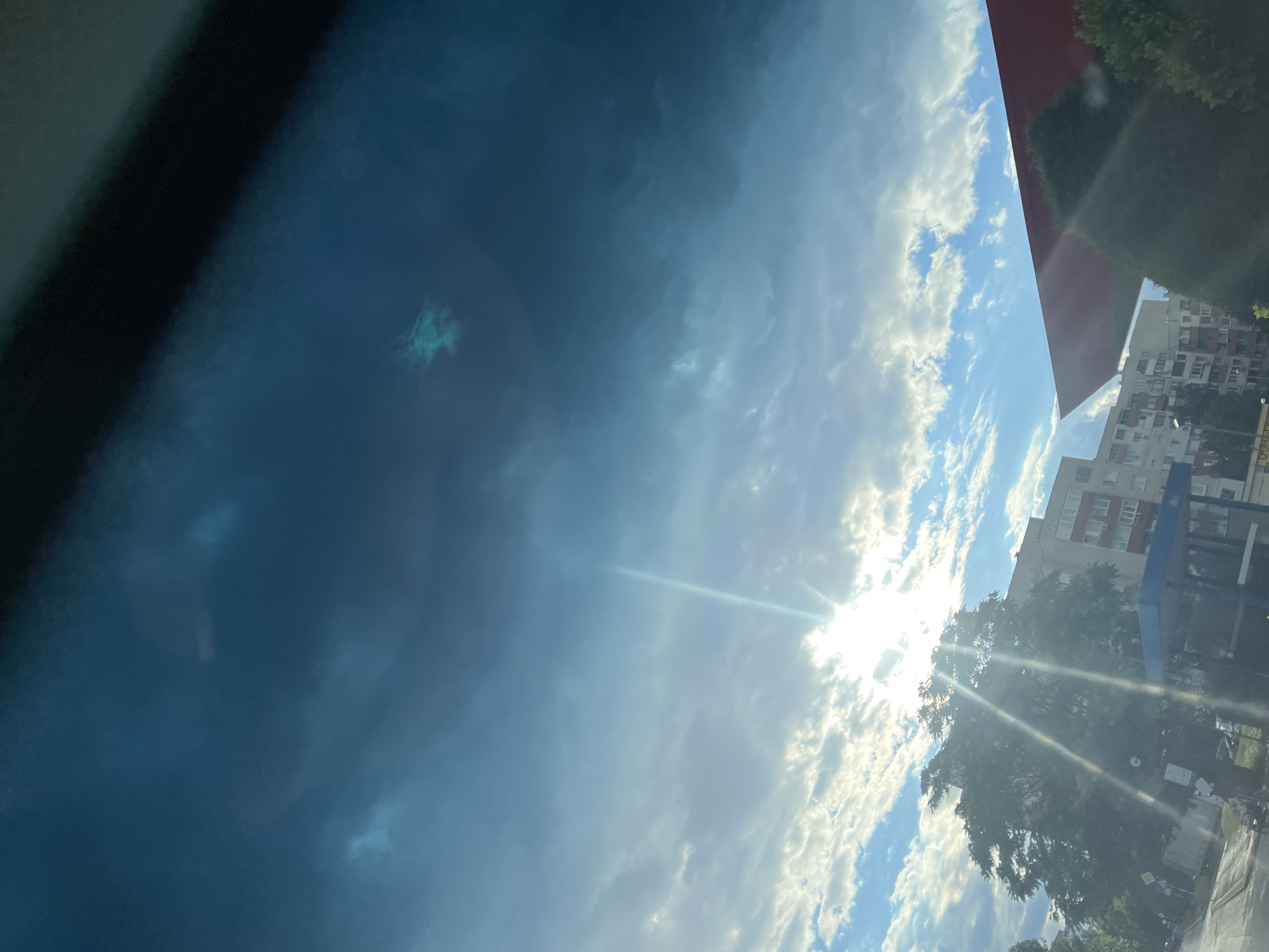The image depicts a sky filled with clouds, with the sun's rays piercing through them. The sun's light creates a lens flare effect, streaking diagonally across the frame. The sky transitions from a bright, cloudy blue at the bottom to a darker, more overcast blue at the top, with some areas appearing almost black. In the lower portion of the image, silhouetted trees and buildings are visible, suggesting an urban environment. A red structure, possibly a roof or canopy, juts into the frame from the right. The scene is captured from a low-angle perspective, looking upwards. There are no people or animals clearly visible in the image.