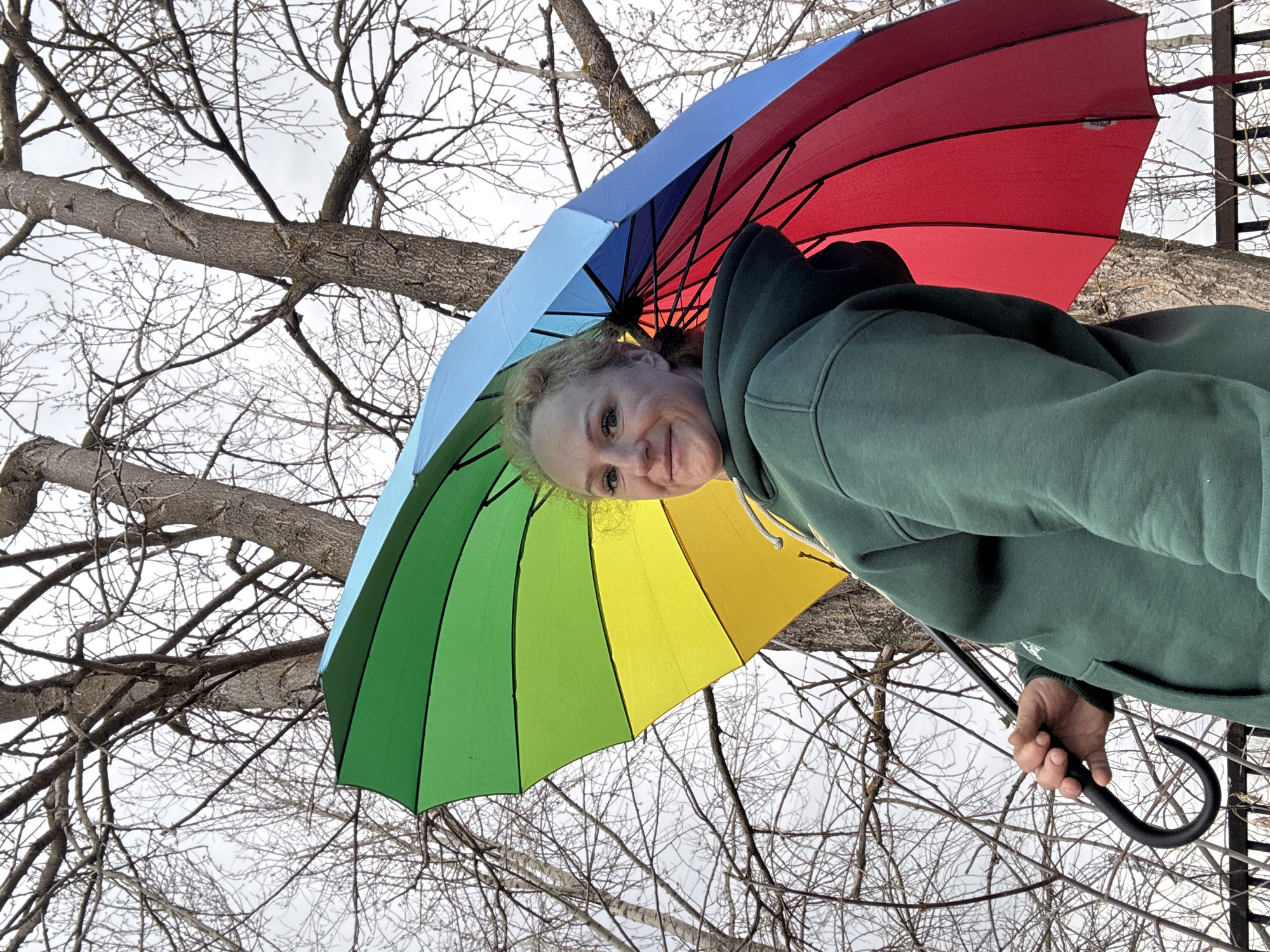A woman in a green hoodie holds a rainbow-colored umbrella in an outdoor setting, likely in Chaikovsky, Russia. Behind her are leafless tree branches against a white sky.