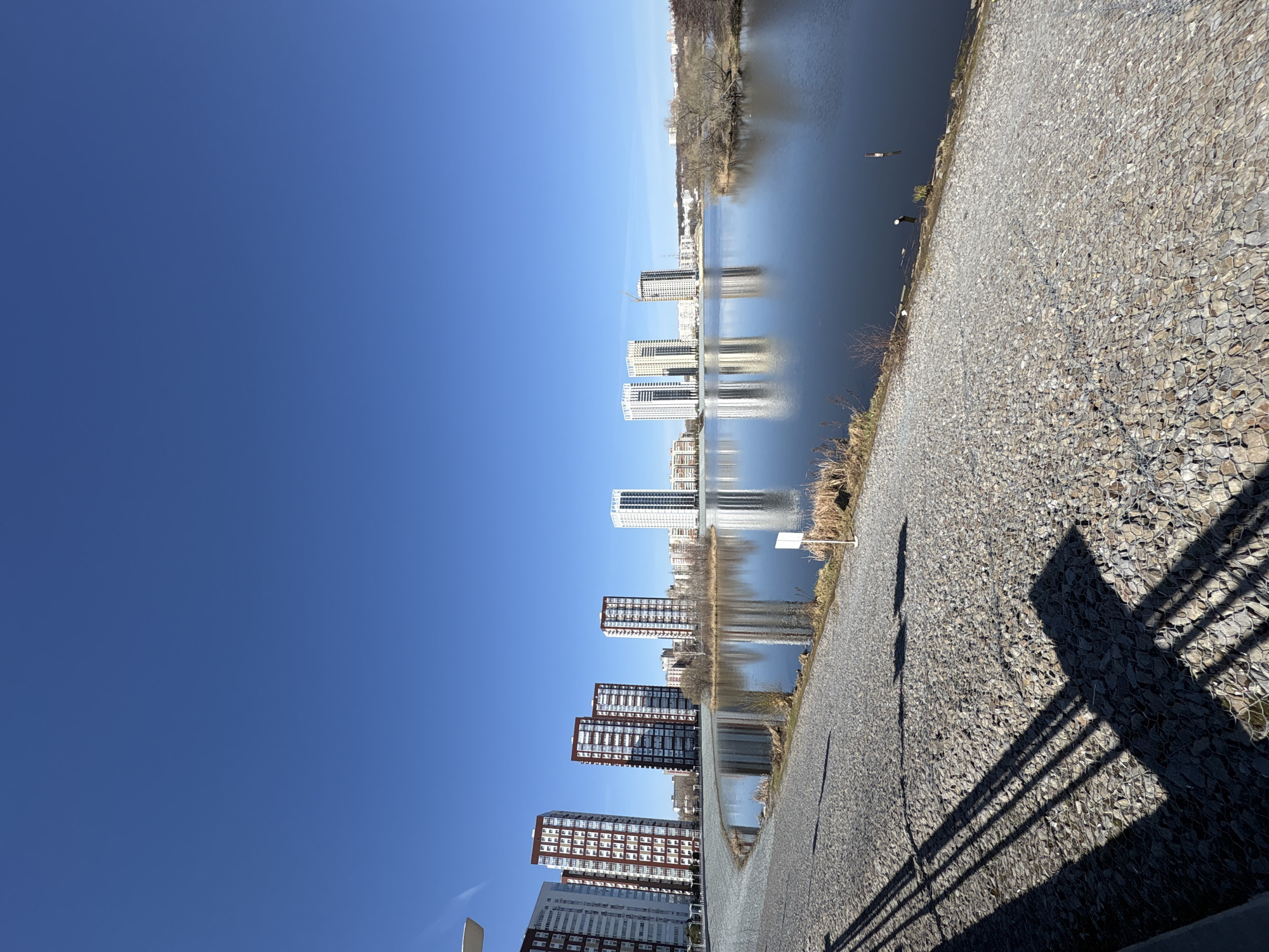 A tilted, wide outdoor shot shows a row of high-rise buildings reflected in a river. The buildings are mostly white or brick-colored. The water is calm with a slight ripple effect. The sky is a clear, bright blue. A pebbled walkway or embankment runs along the right side of the frame, with the shadow of a railing cast upon it. In the background across the river are trees. The location appears to be a city embankment, possibly in Ulyanovsk, Russia. There are no people visible in the frame.