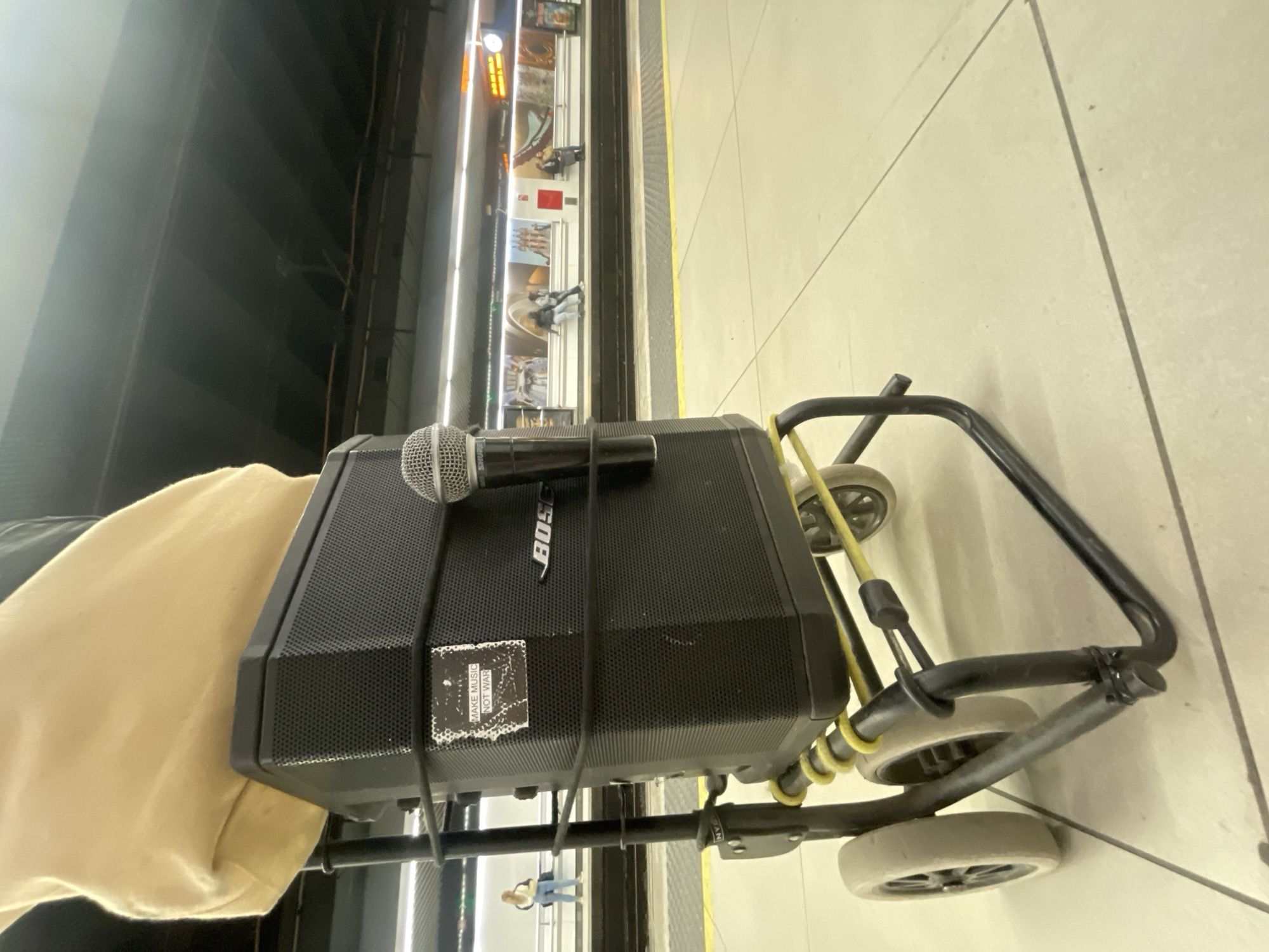 The image shows a Bose portable speaker sitting on a four-wheeled dolly next to a subway platform in Barcelona, Spain. The platform edge features a display of advertisements and posters. A microphone sits on top of the speaker. The scene is captured from an elevated perspective, with a person's arm or shoulder visible near the speaker. People can be seen on the subway platform through the reflection on the speaker.