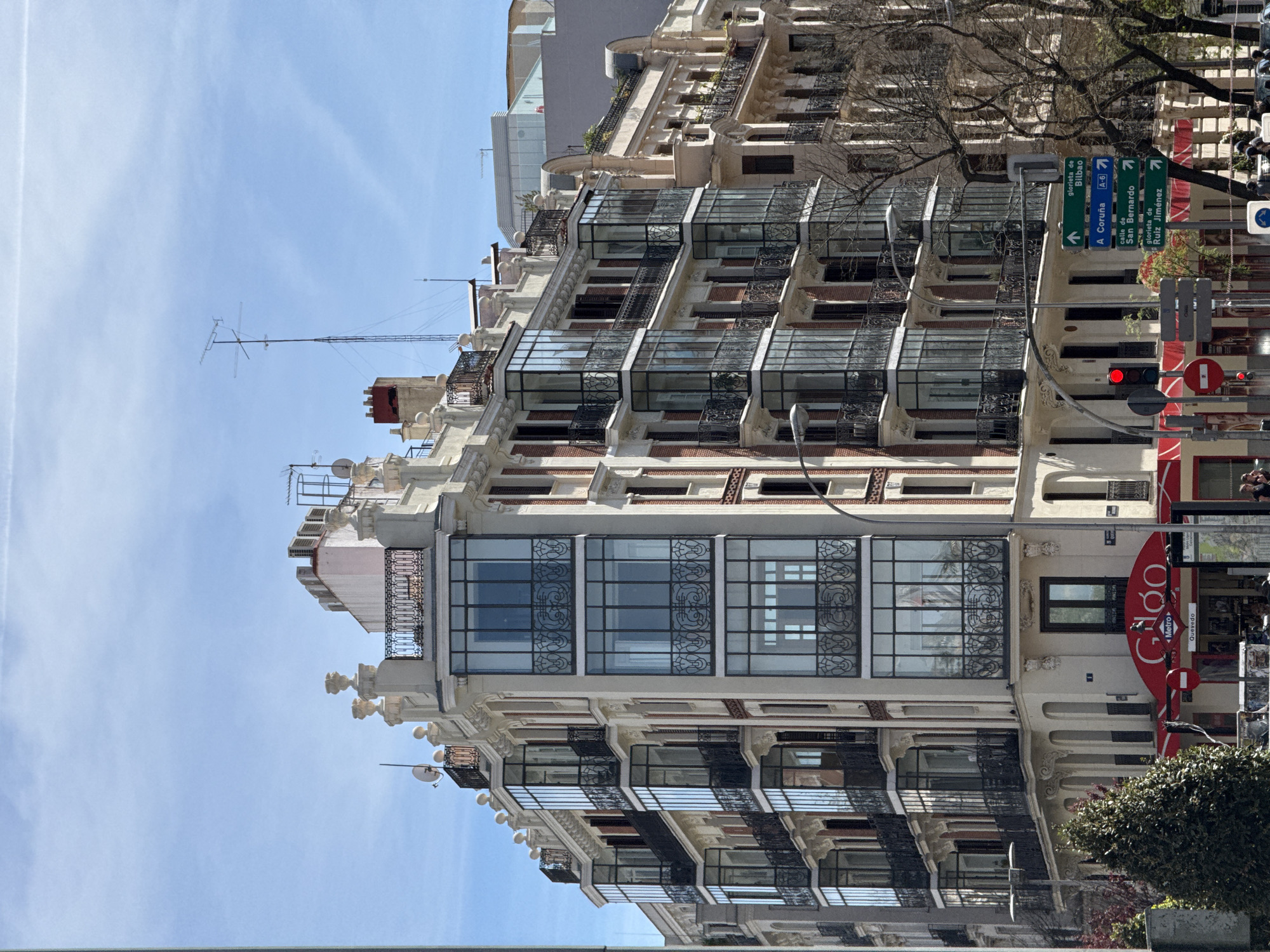 The photograph presents an urban scene in Madrid, Spain, focusing on an ornate building featuring a series of enclosed glass balconies and elaborate architectural details. The facade is predominantly white and is contrasted with dark metal railings and window frames. The building is tall, multi-story, and adorned with various decorative elements.
In the foreground, there is a street with a red traffic light, street signs indicating directions to various locations like Bilbao, A Coruña, and Ruiz Jiménez, and a sign for a "Metro" entrance labeled "Quevedo." There's also a red signage for a brand called "Gido." Some trees and foliage are visible near the bottom of the image.
The sky is blue with some light clouds, suggesting a bright day. The scene appears to be in a busy urban area, potentially a central or well-known district of Madrid.