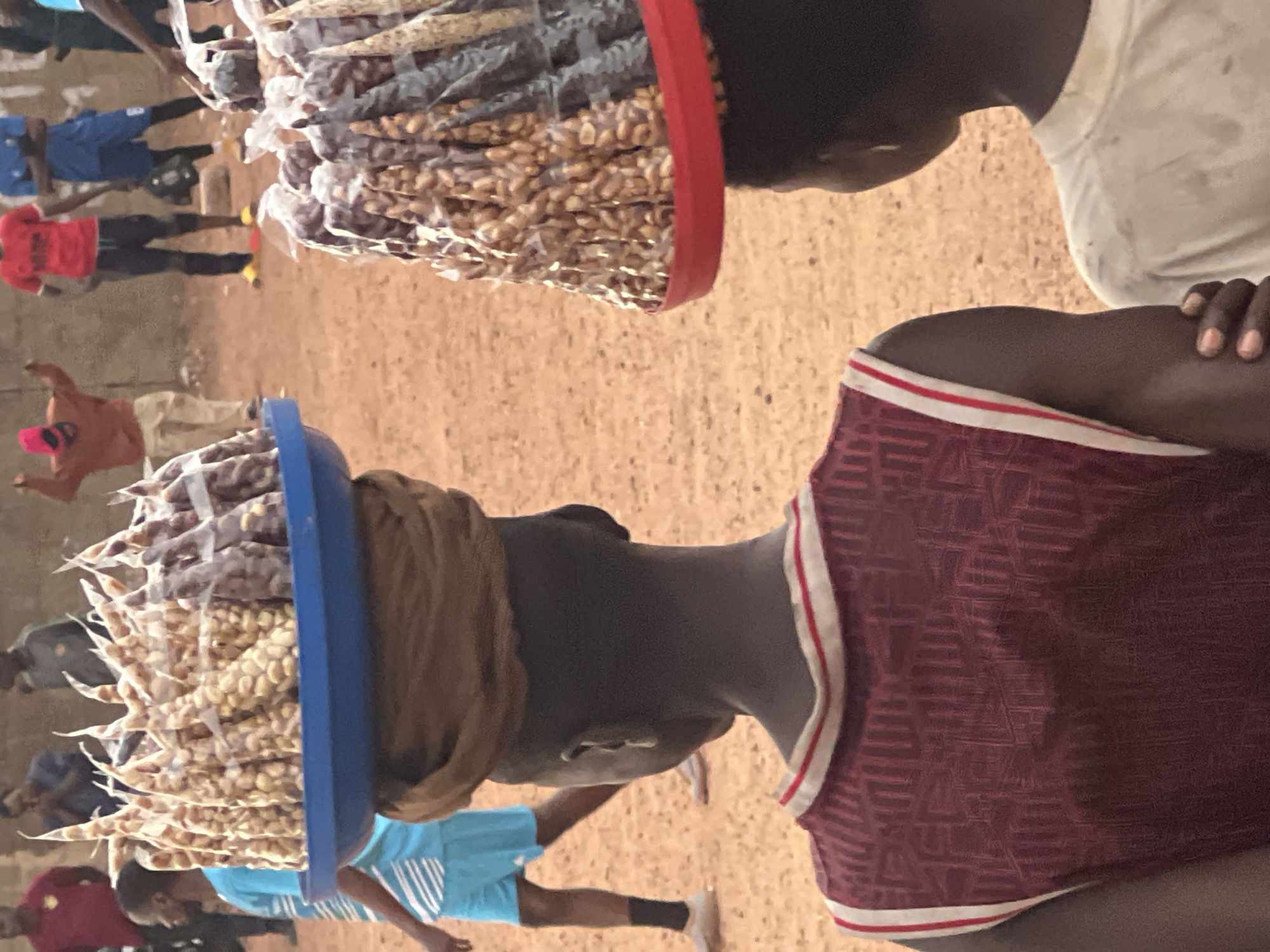 The image shows two people standing close to each other in an outdoor setting. Both individuals have trays balanced on their heads, filled with what appears to be small bags of peanuts. The tray on the head of one person has a blue rim, while the tray on the other's head has a red rim. The ground is a reddish-brown color, and several other people are visible in the background, some wearing blue clothing. The image is likely taken in Jos, Nigeria.