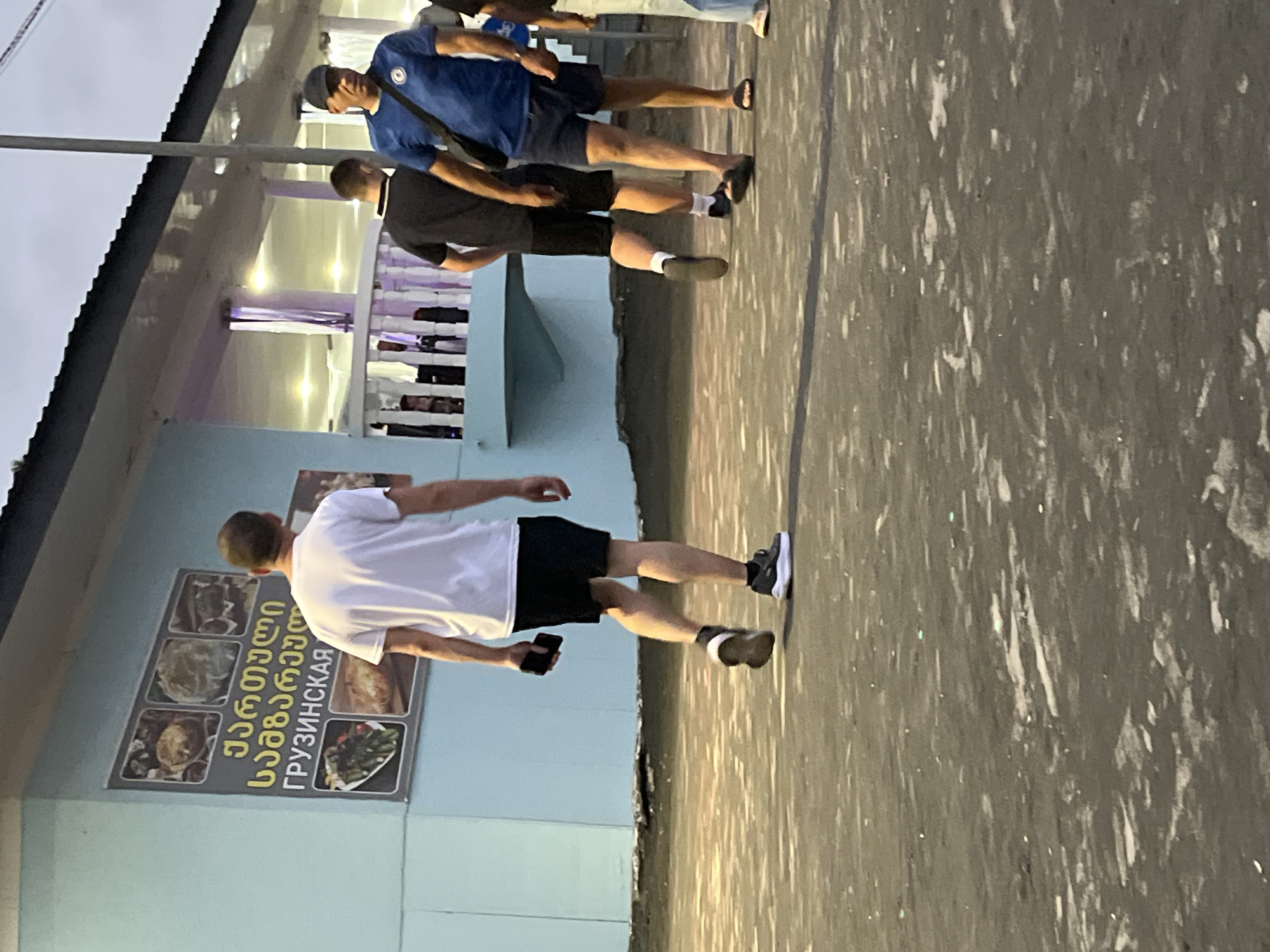 The image depicts a street scene in Ureki, Georgia, with a light blue building on the left featuring a menu board displaying food images and text in Georgian and Russian. Several individuals are present, walking on an asphalt surface. In the foreground, a man in a white t-shirt and black shorts walks away from the camera, holding a phone in his right hand. To his right, two men walk in the same direction, one in a black t-shirt and black shorts, and another in a blue t-shirt and navy shorts, wearing a baseball cap. Further to the right, other individuals are partially visible. The environment suggests an outdoor setting, possibly near a restaurant or establishment, with artificial lighting visible on the building. The sky is overcast, indicating a twilight or cloudy day.