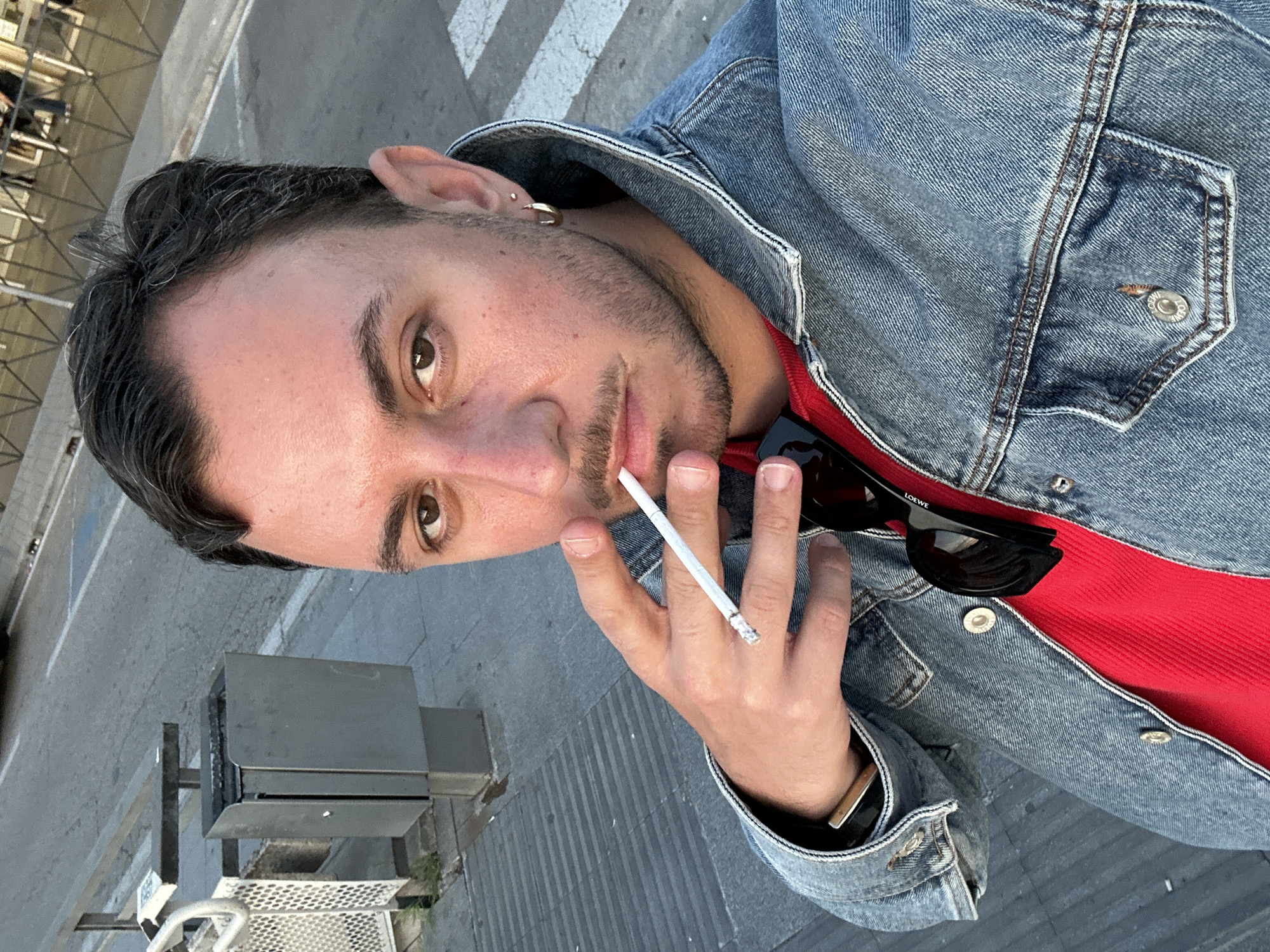 A man is holding a lit cigarette in his right hand. He has short, dark hair and brown eyes. He is wearing a blue denim jacket over a red shirt. A pair of black sunglasses with the brand "Loewe" visible are tucked inside his jacket. He has a gold hoop earring in his left ear. He has stubble on his face. He is standing on a city street, with a pedestrian crosswalk visible in the background. There is a metal trash can or refuse bin next to him. The location is likely Madrid, Spain, given the urban environment.