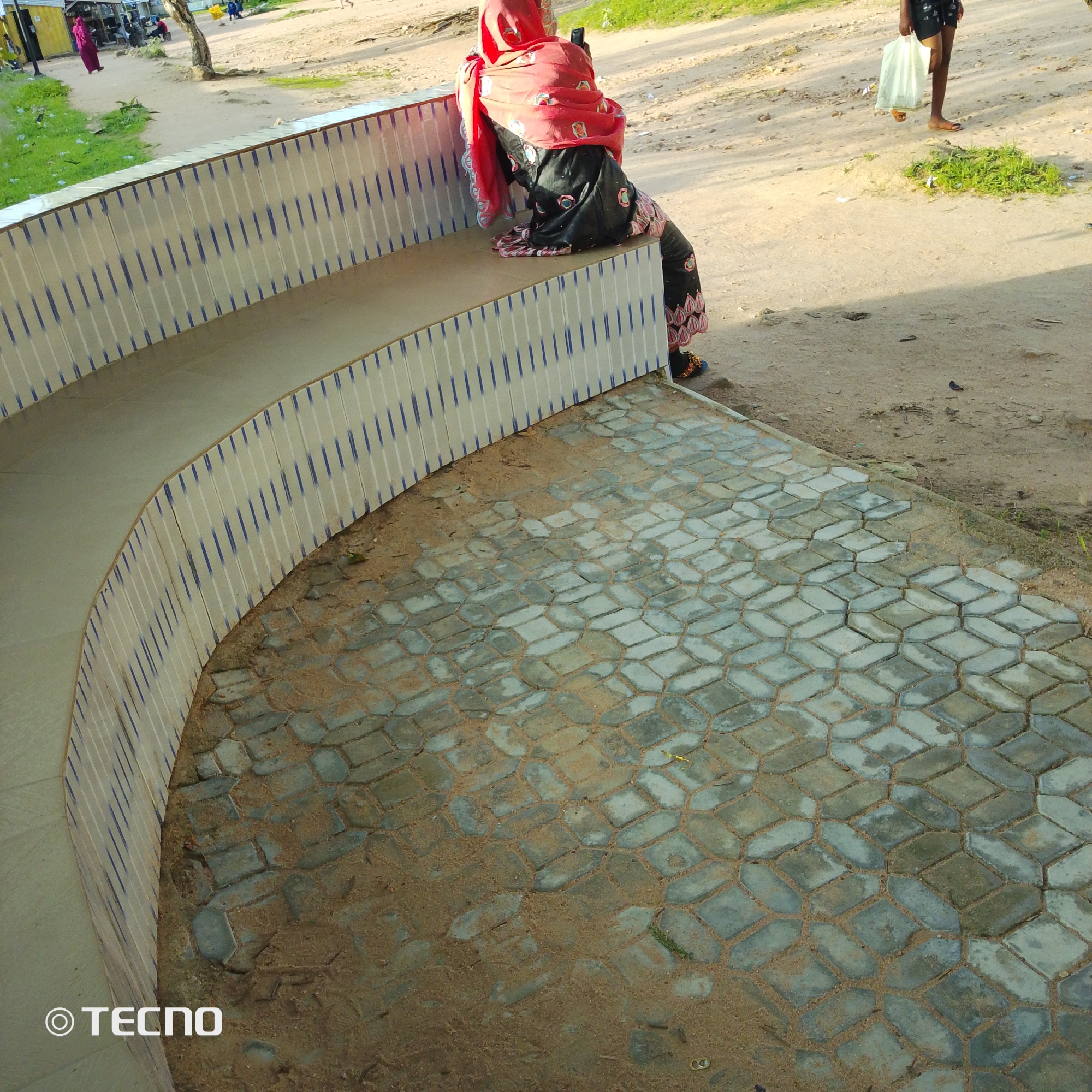 In Jalingo, Nigeria, a woman wearing a red hijab and a black garment with colorful embroidery is seated on a tiered, curved bench with blue and white striped tile accents. The bench is adjacent to a paved area with interlocking hexagonal concrete pavers interspersed with sand. In the background, a dirt path leads to buildings and sparse vegetation. Another person, partially visible, is walking on the path carrying a light-colored plastic bag. The scene is lit by natural daylight, casting shadows on the ground. The "TECNO" logo is visible in the bottom left corner of the image.
