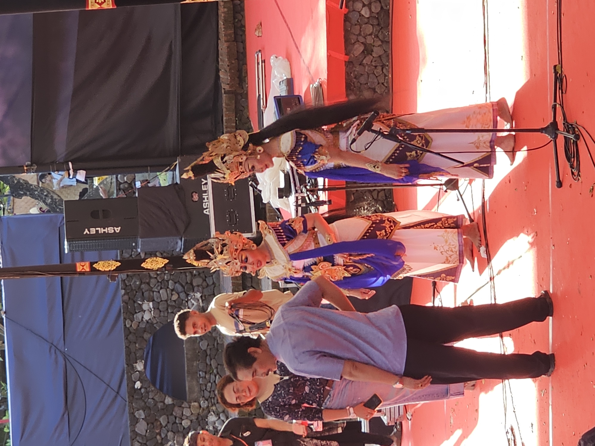 The image depicts an outdoor cultural performance in Denpasar, Indonesia. The scene is set on a bright red carpet stage, with a stone wall and foliage in the background. Two female performers in elaborate traditional Balinese costumes, featuring gold headdresses and ornate blue and white attire, are the central focus. They stand on the stage, with one looking towards the right and the other facing left. A man in a light blue shirt and dark trousers stands between the performers and the camera, interacting with them. Another man in a tan shirt is visible behind them, holding a microphone cable. Several audio speakers, branded "ASHLEY," are positioned behind the performers. The overall atmosphere suggests a formal cultural event or festival.