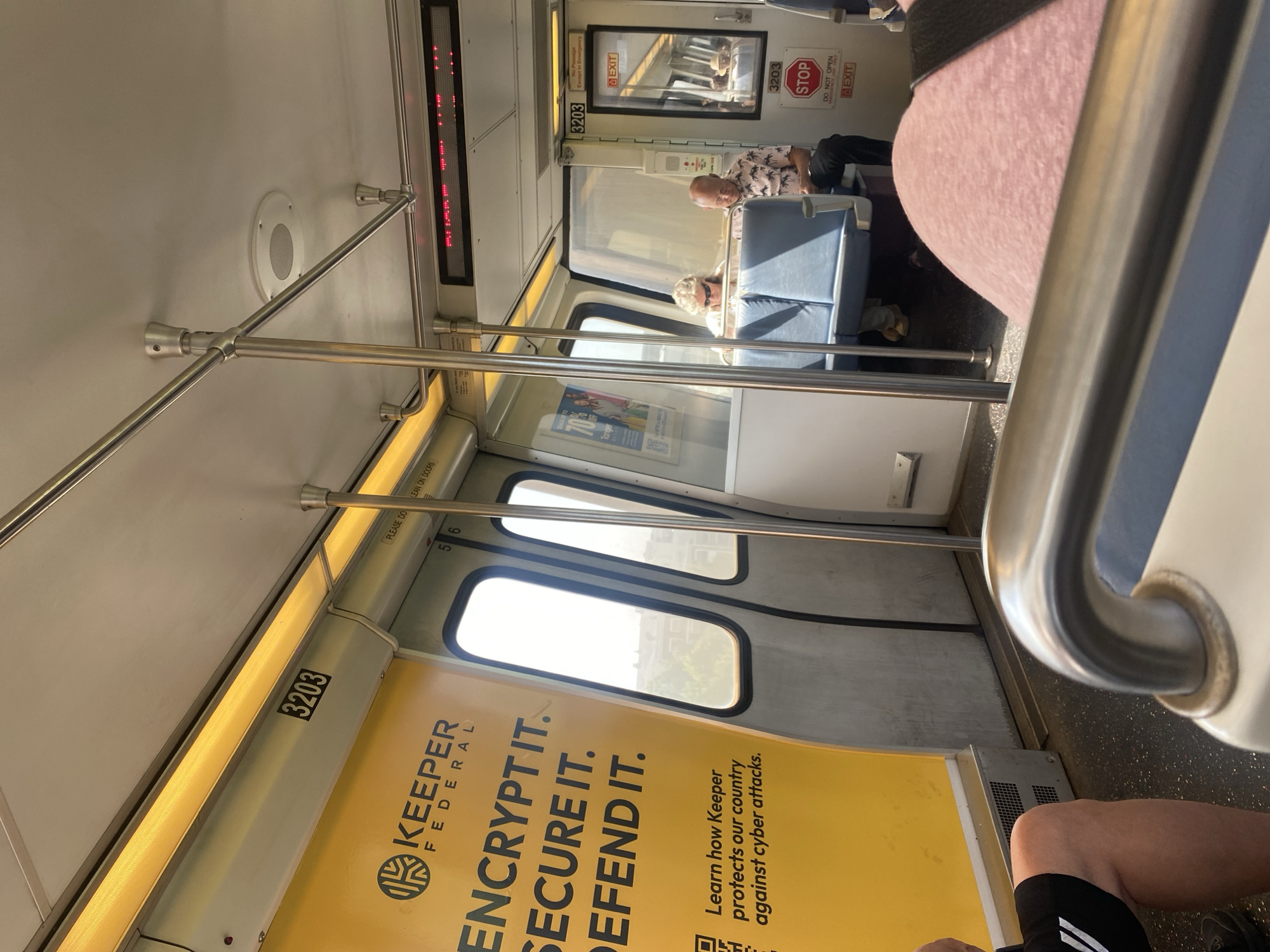 This media file depicts the interior of a train car, likely in Washington, USA. The perspective is from a seated passenger looking forward. Two people are visible: a man with a patterned shirt and a woman with sunglasses, both seated. The train appears to be in motion or stopped, judging by the daylight visible through the windows and the presence of passengers.
Prominent objects include handrails, seats, windows, and advertisements. A large yellow advertisement for "KEEPER FEDERAL" with the slogan "ENCRYPT IT. SECURE IT. DEFEND IT." is featured prominently. Another ad displays "DEALS UP TO 70% TONGER". Signs indicating "STOP", "EXIT", and "DO NOT OPEN" are also visible, along with the car number "3203".
The lighting suggests it is daytime. The overall mood is casual and mundane, typical of public transportation. The scene is a snapshot of daily life on a train.