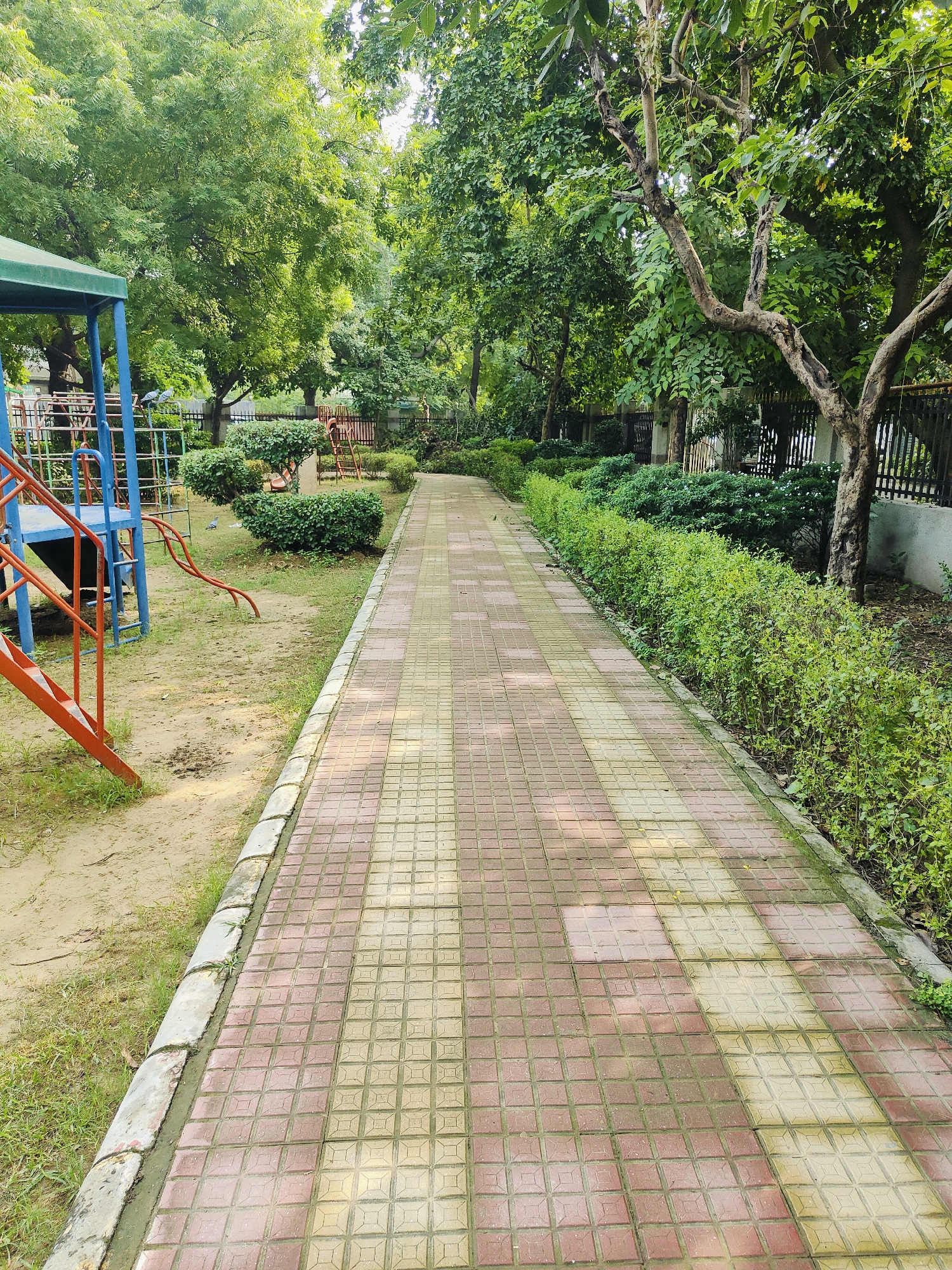 The image depicts a park in Gurgaon, India. A paved pathway, made of red and yellow square tiles, runs diagonally from the foreground to the background on the right side of the image. To the left of the path is a children's playground with blue and orange metal structures, including a slide and steps. Beyond the playground, there are manicured bushes and trees. A black metal fence borders the park on the right side, behind which are more trees and some structures. The ground in the park is a mix of sand, dirt, and grass. Sunlight filters through the lush green foliage, creating dappled lighting on the path and playground. The overall scene suggests a well-maintained urban park.