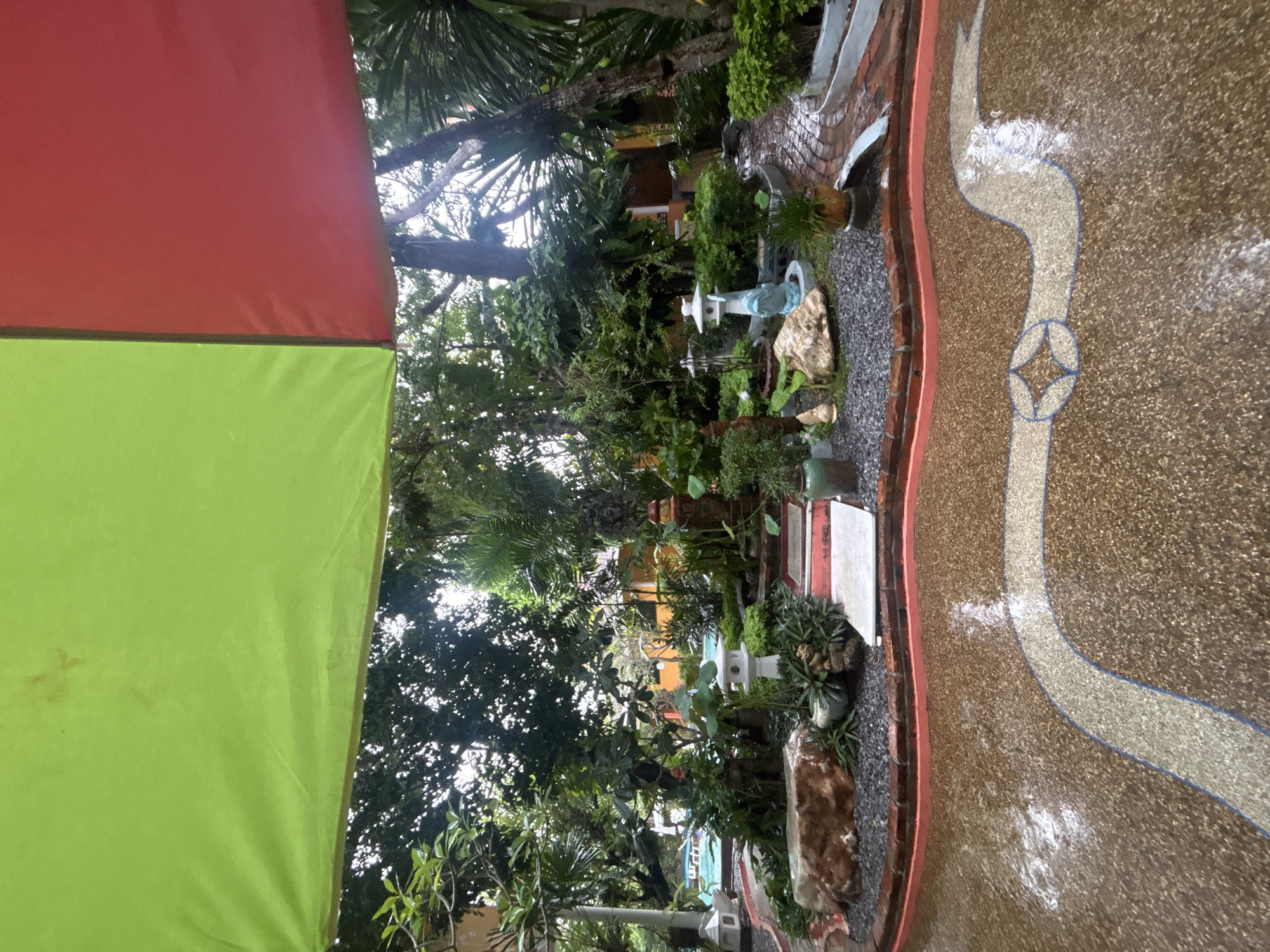 This is a photograph taken from a low-angle perspective looking up at a garden area, possibly a backyard or patio, in Chalong, Thailand. The foreground features a wet, speckled ground with a curving decorative inlaid pathway. In the mid-ground, a landscaped garden is visible, characterized by gravel paths, various potted plants, and decorative elements such as stone lanterns and a large rock. A red brick pathway leads into the garden. Beyond the garden, trees with lush green foliage dominate the scene. In the far background, a swimming pool can be partially seen, and buildings with orange facades are visible. The upper portion of the image is covered by a canopy or umbrella, with sections of bright green and red fabric. The overall impression is of a well-maintained outdoor space, likely experienced during or after rainfall, given the wet surfaces. No people are present in the image.