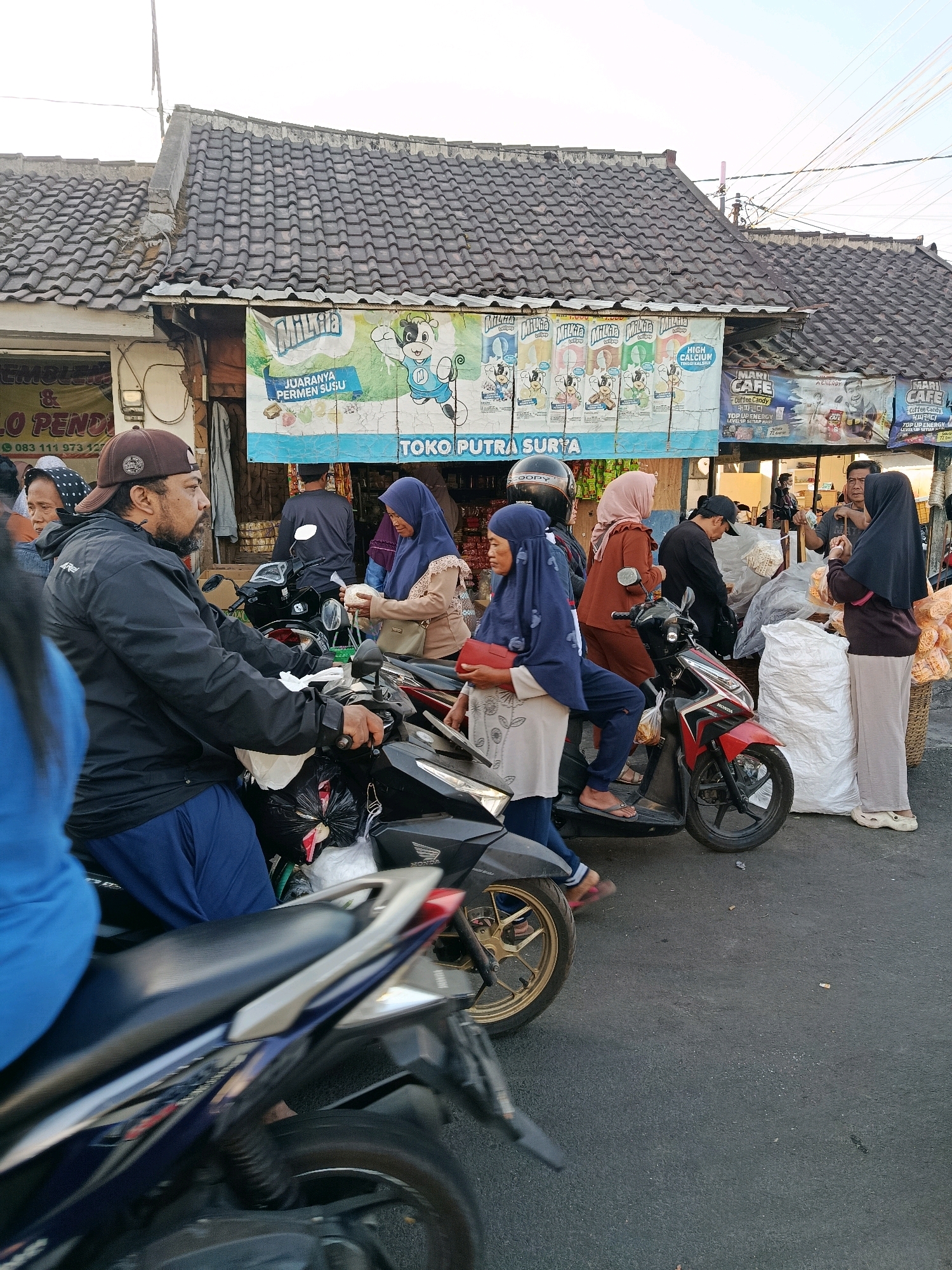 The image depicts a street scene in Bululawang, Indonesia, with several people and motorbikes in front of a small shop. In the foreground, a man wearing a brown cap and black jacket is seated on a motorbike, looking to his right. Beside him, a person in a blue top is partially visible on another motorbike. Behind them, several individuals are interacting. A woman in a blue hijab and light-colored top is holding a red bag. Another woman in a rust-colored hijab and matching outfit is standing next to her. A person in a blue hijab and a light grey shirt is also present. In the background, a shop displays banners for "TOKO PUTRA SURYA" and various products. A man in a grey shirt is seen near the shop entrance, and another person in a black hijab is standing to his right. Motorbikes are parked and in use, with one red and black motorbike prominently featured with a woman in a blue hijab and a grey shirt holding a white object. The ground is paved, and the sky is overcast. The environment suggests a busy marketplace or street-side gathering.