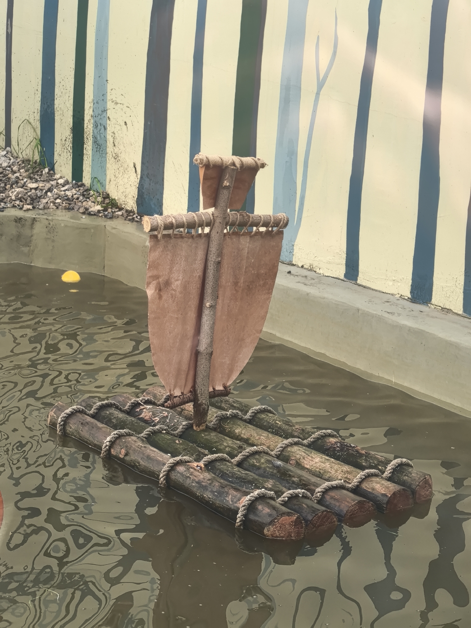 A small, rustic raft sits in murky water. The raft is made of several logs lashed together with rope. A makeshift mast rises from the center of the raft, with a sail attached. The water reflects the raft and the surrounding environment, creating a rippled effect. A yellow ball floats in the water behind the raft. The background is a wall painted with vertical stripes in shades of green, blue, and beige. A pile of gravel is visible at the base of the wall. The scene is an artificial environment, likely part of an exhibit or display. This image was taken at the "Gorodskaya Gorka" park in Perm, Russia.