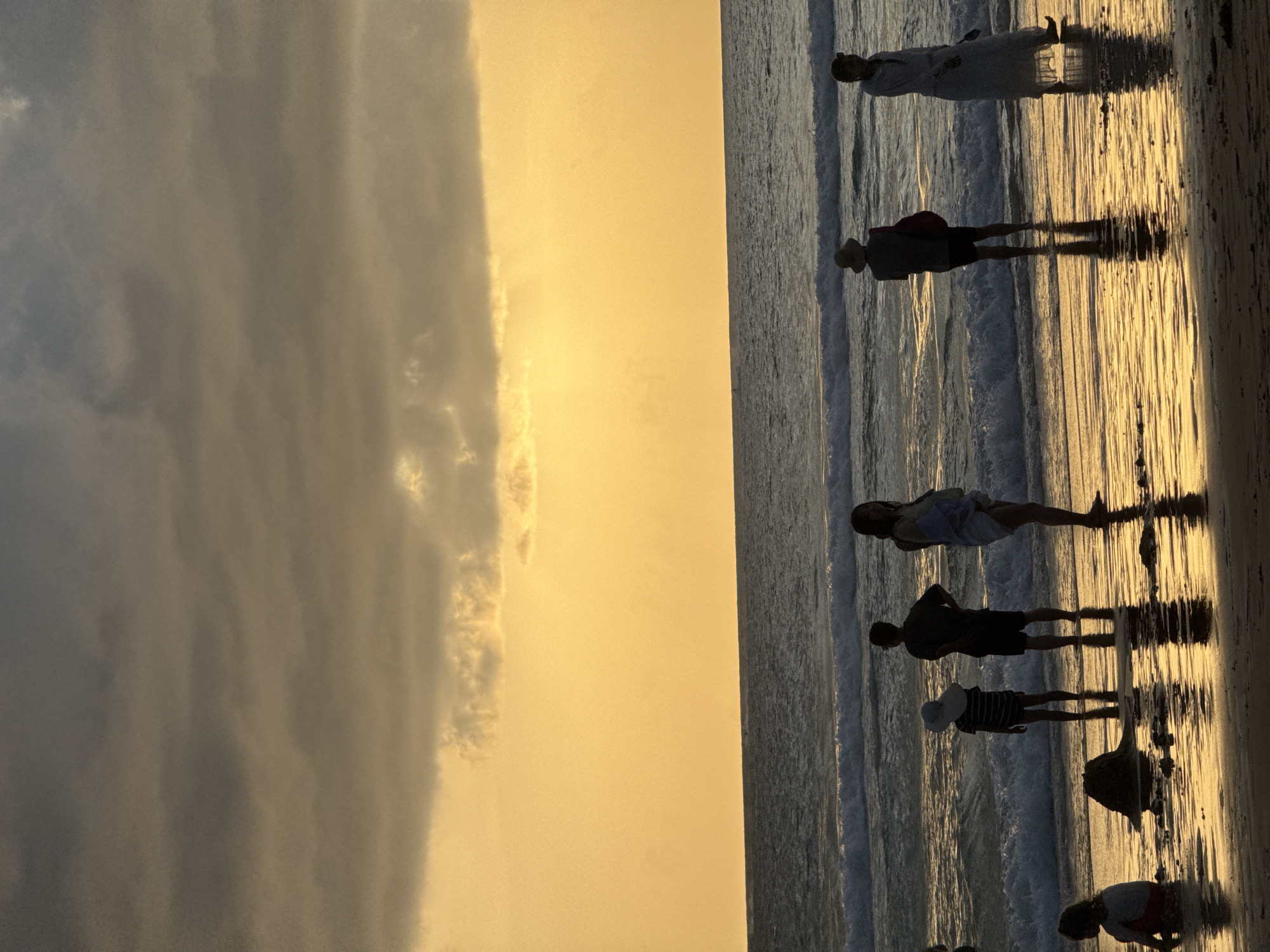 The image depicts a beach scene at Karon Beach, Thailand, during sunset. The sky is filled with warm yellow and orange hues, with dark clouds obscuring the upper portion. The ocean is a deep blue with gentle waves breaking on the shore. Several people are visible in the shallow water and on the wet sand, silhouetted against the bright sunset. On the left, a child sits in the water, and a sandcastle is to their right. Further to the right, a family stands in the water; a child in a striped shirt and a wide-brimmed hat faces away from the camera, a man in dark shorts stands next to them, and a woman in a light-colored dress stands further right. Towards the right side of the frame, two more figures are visible, one wearing a hat and the other in a long white dress. The reflections of the people and the sky are visible on the wet sand. The overall atmosphere is serene and picturesque.