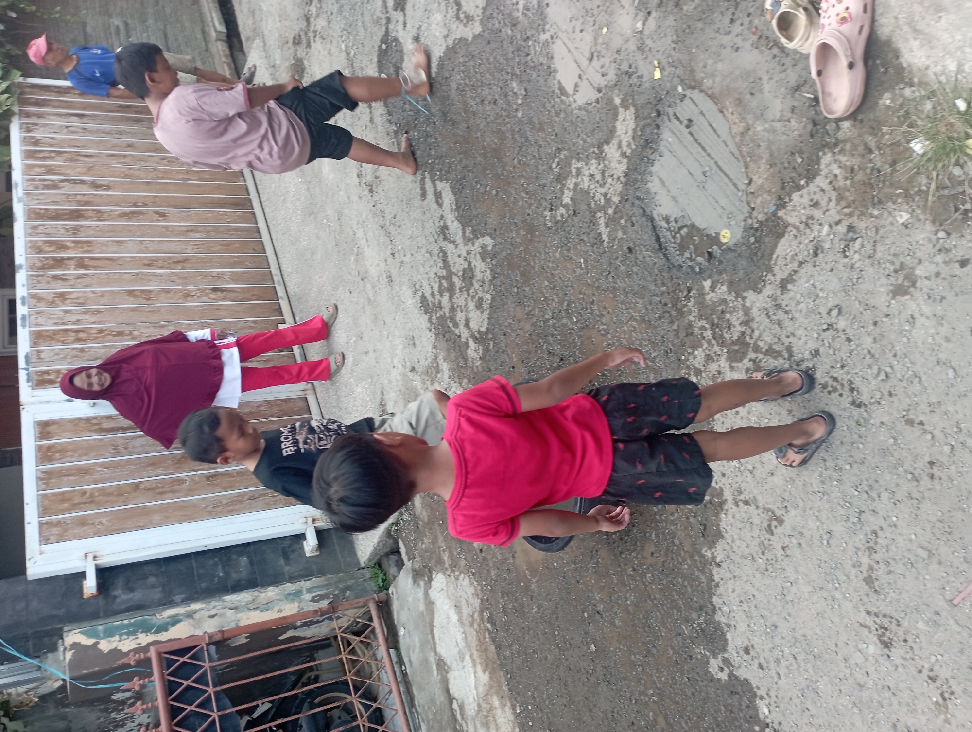 In an outdoor, urban environment, a group of people are visible. A woman wearing a dark red hijab and pink pants stands near a wooden gate. To her right, a boy in a light brown t-shirt and dark shorts is facing away from the camera, looking down. A younger boy in a red t-shirt and black patterned shorts is in the foreground, also facing away. Another boy, wearing a dark t-shirt with a white graphic, is to the left of the woman. An older man in a blue shirt and light shorts, wearing a pink cap, stands further to the right. The ground is a mixture of dirt and gravel, with some puddles and a circular manhole cover. There are also a pair of tan clogs and a pair of pink crocs on the ground. The scene appears to be an informal gathering or play area.