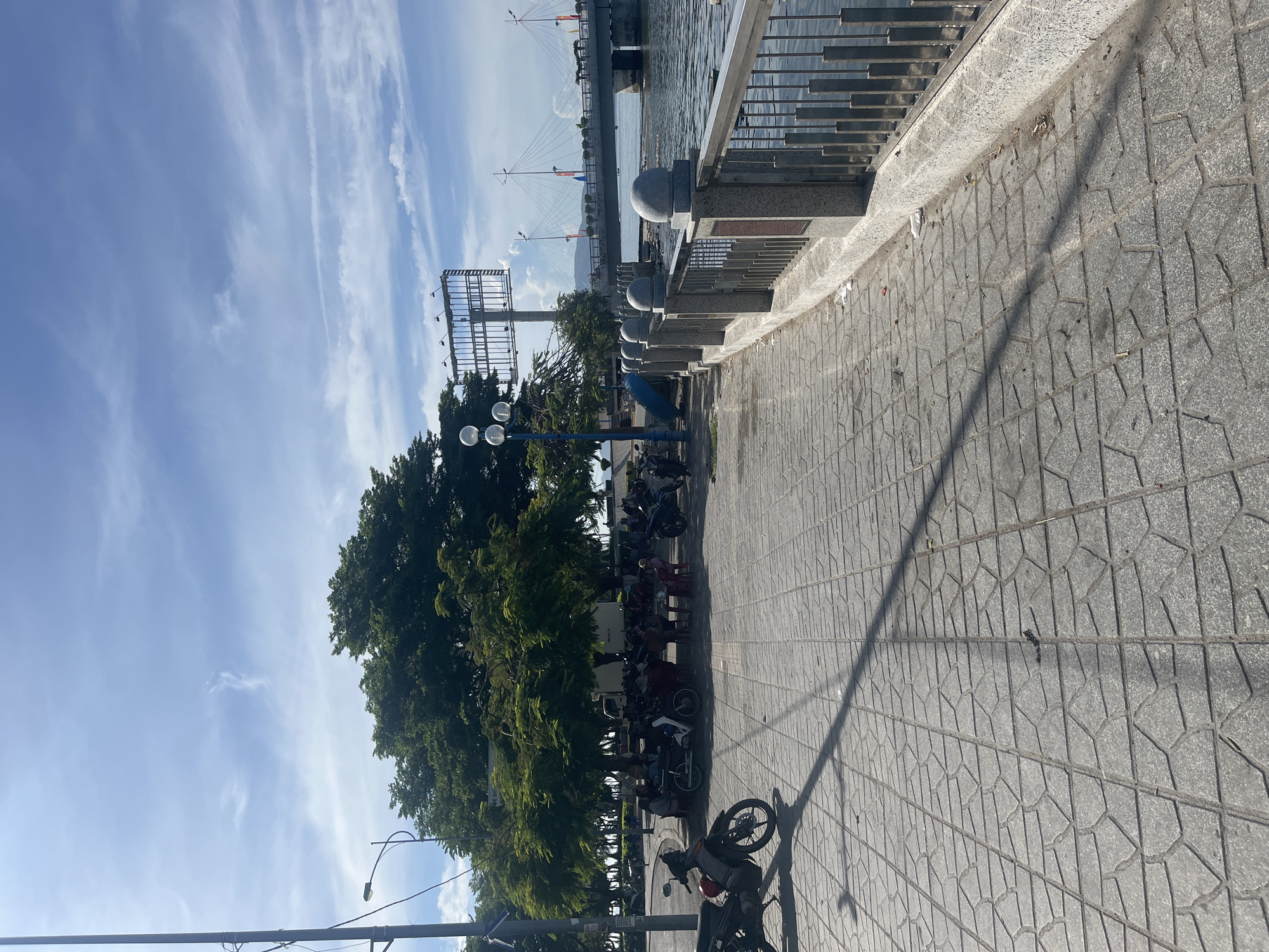 The image depicts a waterfront view in Nha Trang Ward, Vietnam, characterized by a paved walkway lined with a decorative railing along the water's edge. In the background, a cable-stayed bridge is visible. The walkway is made of interlocking geometric-patterned gray pavers. Trees line the left side of the walkway, with parked motorcycles and scooters clustered beneath them. Streetlights are interspersed along the walkway. The sky is partially cloudy, with blue patches. A billboard-like structure stands on the left. The sun is casting shadows across the walkway.