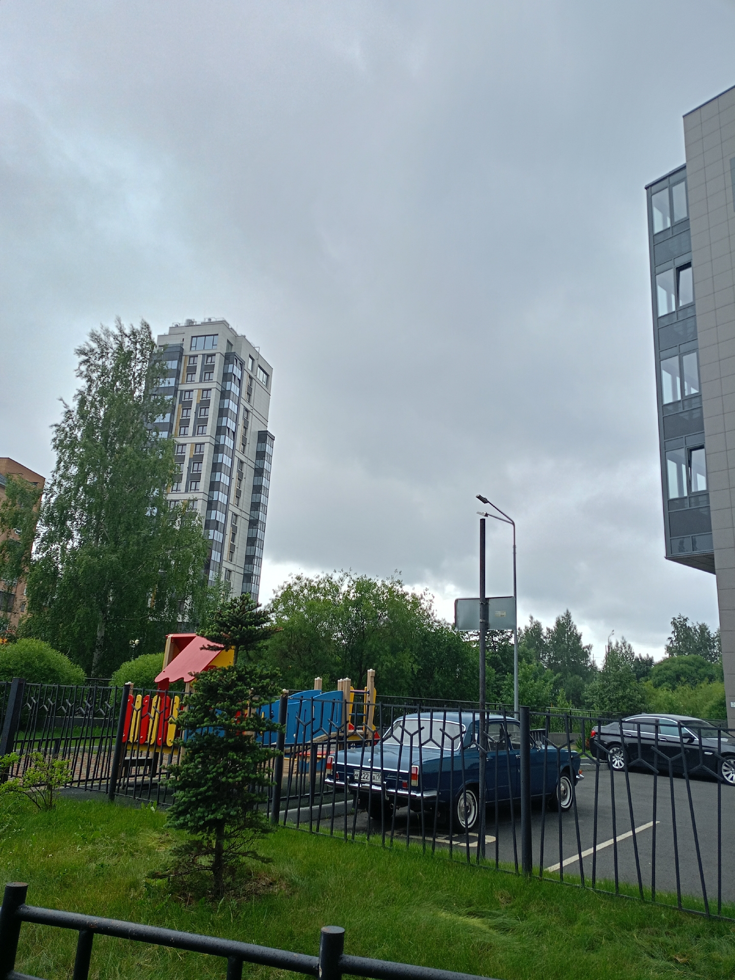 This media file captures an outdoor scene on a cloudy day. The primary subjects are two cars parked on an asphalt surface behind a black metal fence. The car in the foreground is a dark blue vintage sedan with whitewall tires. Behind it, a black modern sedan is visible, partially obscured by the fence.
Beyond the fence, a children's playground with colorful structures in red, yellow, and blue is set against a backdrop of lush green trees. A tall, modern apartment building with a white and grey facade and numerous windows stands to the left of the frame, partially covered by a large, leafy green tree. To the right, a section of another modern building, also with grey cladding and large windows, is visible. A street lamp with a sign beneath it is positioned between the playground and the buildings.
The sky is overcast, indicating a cloudy day, possibly with overcast conditions. The presence of a playground suggests a residential area, likely an apartment complex. There is no discernible activity taking place; the scene appears to be a still moment in time.
Visible text includes license plates on the cars, though the characters are difficult to read clearly due to the image resolution. One license plate reads "K 22 A10". The overall mood is neutral, set by the overcast weather and the mundane arrangement of objects.