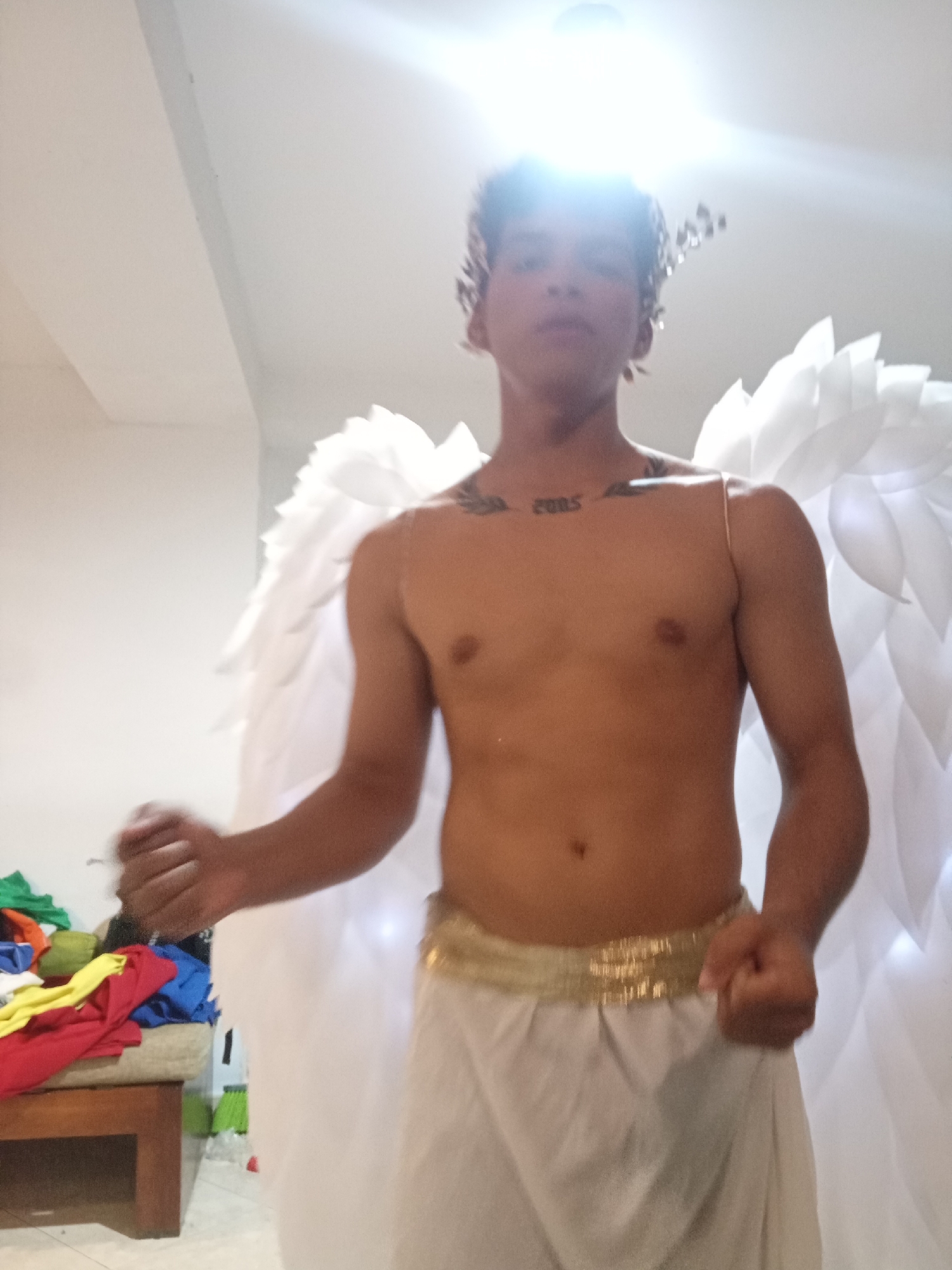 A shirtless person wearing a white toga, a gold belt, and large white wings stands in a brightly lit room. The person also wears a headdress of some kind. A cluttered bench with colorful clothing is visible in the background. The location is likely indoors, but no details suggest it is specifically in the city of Playa El Agua, Venezuela.