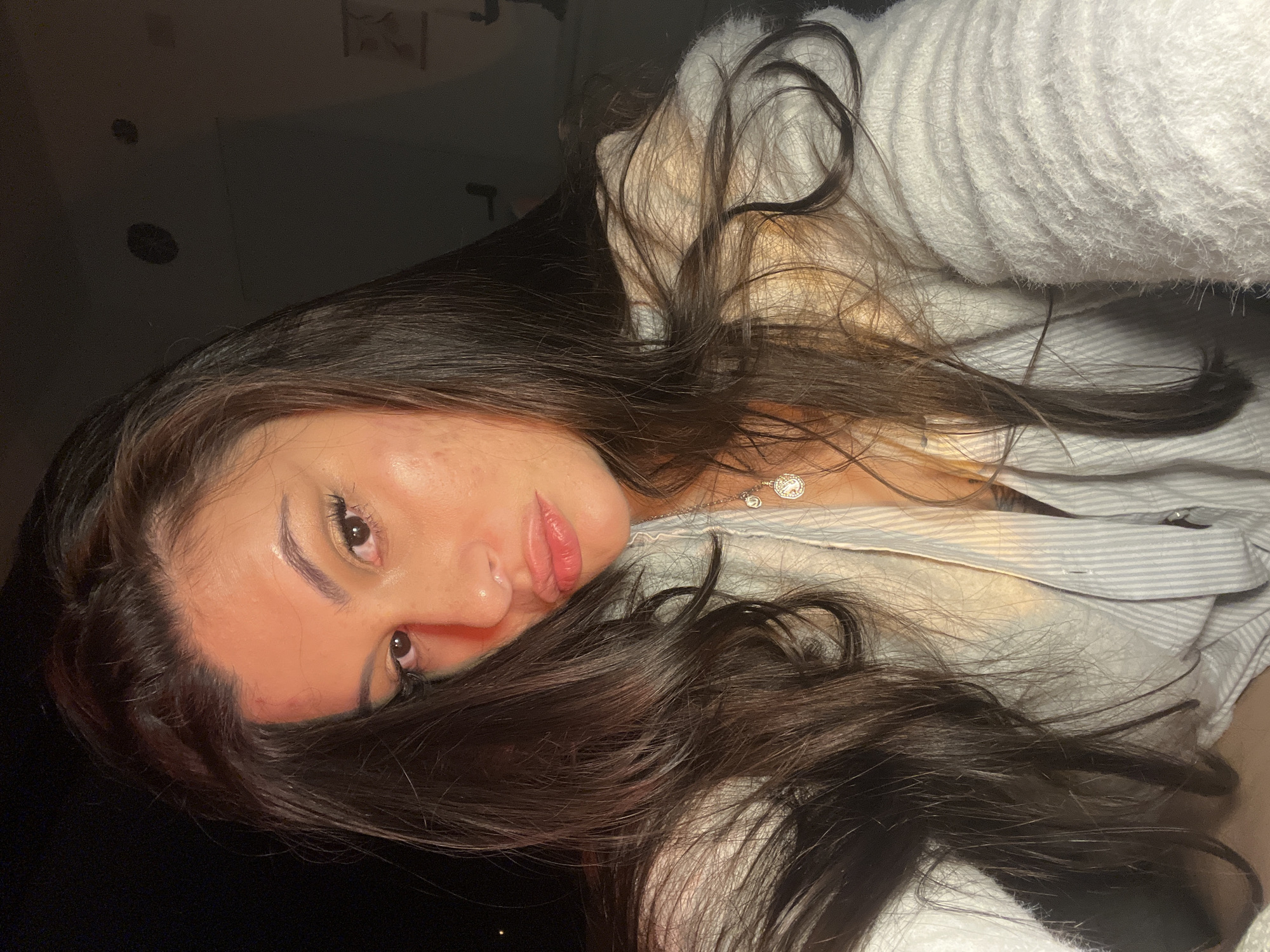 A close-up portrait features an adult female with long, dark, wavy hair, parted on the left, cascading over her shoulders and chest. She looks directly at the camera, her face partially illuminated by a strong, warm, orange-red light source from the left, which casts a reddish hue on her left cheek and forehead. The right side of her face receives softer illumination. Her full lips are slightly pursed, and makeup, including eyeliner and lip gloss, is present. Minor skin texture and possible blemishes are visible on her cheeks and forehead. She is wearing a light-colored, textured, possibly fuzzy knit top or sweater over a lighter, vertically striped button-up shirt, which is partially unbuttoned. A silver-colored chain necklace with two distinct pendants, one oval and one circular, both exhibiting intricate designs, is visible around her neck. The background is predominantly dark and out of focus, with indistinct, blurry shapes faintly discernible on a wall to the far right, indicating an indoor environment with low ambient light or night conditions. The scene is located in Kiryat Bialik, Israel.
