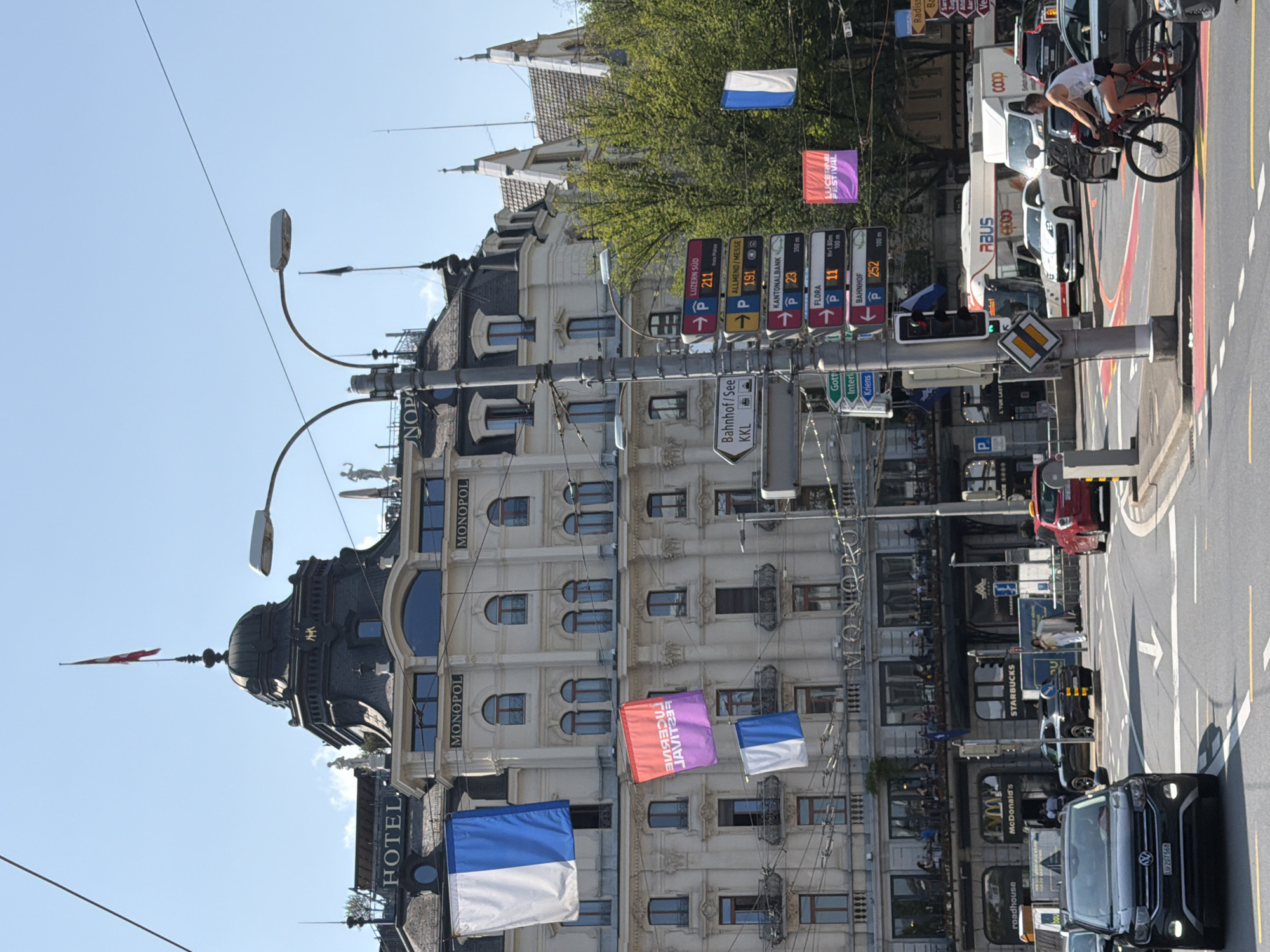 In an urban environment on a clear day, a large, ornate hotel building with a domed roof and a flag atop it dominates the background. Multiple banners hang from the hotel facade, displaying colors and text, including a red and purple banner that reads "LUCERNE FESTIVAL". A street lamp with multiple signs is positioned in the foreground, providing directions to various locations such as "Luzern Sud", "Allmend/Messe", "Kantonalbank", "Flora", and "Bahnhof". Several cars, including a black SUV and a red sedan, are on the street. A man on a bicycle rides past the street lamp. Other businesses like McDonald's and Starbucks are visible in the lower portion of the image. The scene depicts a street intersection with pedestrian walkways and road markings.