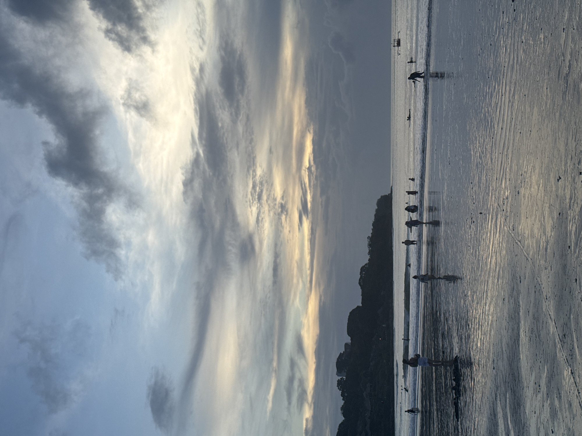 The image captures a serene beach scene during either sunrise or sunset, characterized by its dramatic sky and silhouetted figures. The upper portion of the frame is dominated by a cloudy sky, displaying a mix of deep blues, soft grays, and warm golden light breaking through, suggesting the sun is low on the horizon. This light creates striking streaks and contrasts against the darker cloud formations.
The lower half of the image features a wide expanse of wet sand and shallow water, which brilliantly reflects the colors of the sky and the lingering light. The wet sand exhibits subtle patterns and ripples, indicating the tide's movement. Several people are visible as dark silhouettes against the reflective surface. They are scattered along the shoreline, some wading in the shallow water, others walking on the sand. Their activities appear leisurely, possibly enjoying a stroll, playing, or searching for shells. One individual is bent over, looking at the ground, while another stands near what might be a small boat or raft. Further out in the water, a small, square-shaped object, possibly a buoy or a marker, is faintly discernible.
On the right side of the image, a dark, treelined landmass or distant hill forms a silhouette against the horizon, grounding the vastness of the sky and water. The overall scene conveys a sense of tranquility and natural beauty, typical of the golden hours at a beach. No text is visible in the media file.
