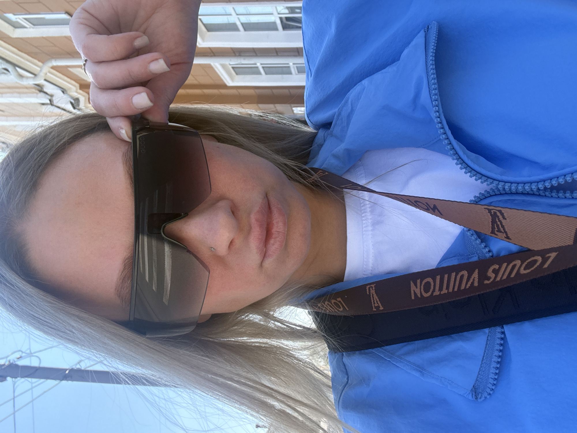 A woman with fair skin and light blond hair is posing for a selfie outdoors. She is wearing large, dark sunglasses and a blue zippered jacket. A Louis Vuitton strap or belt is visible across her chest. She has a small stud piercing on her nostril. She is holding her sunglasses with her left hand, which has a ring on the ring finger. The background shows an urban setting with a light-colored building and a partially visible blue sky. Power lines are slightly visible in the lower left corner. The photograph was taken in Perm, Russia.