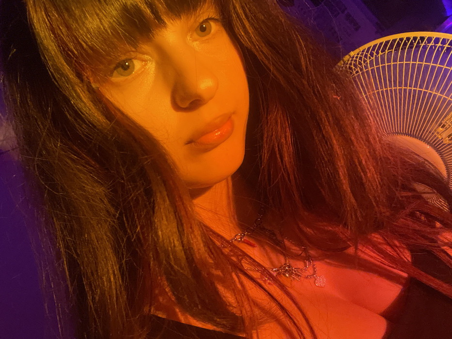 A woman with dark hair and bangs looks directly at the camera. She is wearing a silver necklace. A fan is visible behind her to the right. The lighting is warm, with a strong orange tint across her face and hair, while the background has a blueish tint.