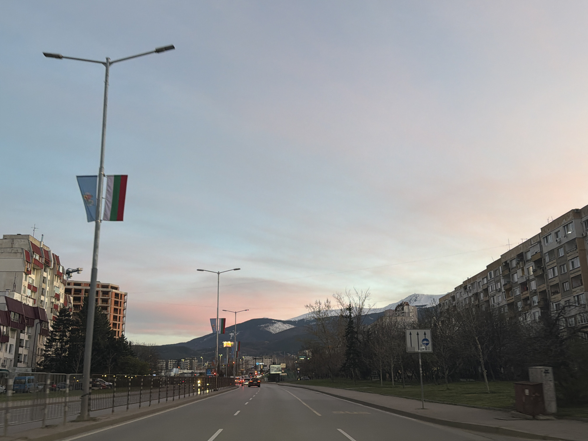 The image shows a street in Sofia, Bulgaria, at dusk. The road is straight, with two lanes in each direction, and white lane markings. Cars are visible in the distance. On the left side of the road, there is a metal guardrail, followed by trees and buildings, including a building under construction. A lamppost displays two flags, one of Sofia and one of Bulgaria. On the right side of the road, there is a grass strip, trees, and a large residential building. In the background, a mountain range is visible, with snow-capped peaks. A road sign indicates lane directions. The sky is a mix of light blue, white, and pink.