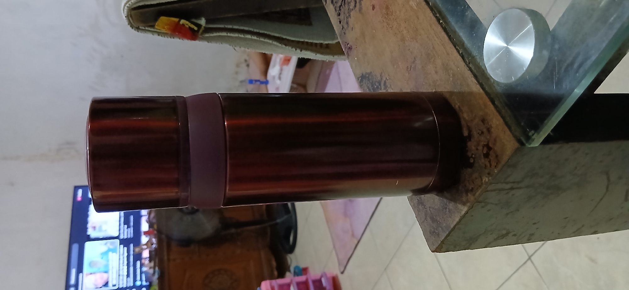 The image displays a metallic maroon-colored thermos bottle standing upright on a dark, textured surface that appears to be the top of a piece of furniture, possibly a table. The thermos has a darker maroon cap and a lighter maroon band around its middle. In the background, a television screen is visible, displaying what appears to be a news program or streaming content with people's faces. To the left of the television, a wooden cabinet or dresser is partially visible. A black fan stands behind the cabinet. Further to the left, a pink plastic organizer with multiple drawers is also present. The floor appears to be tiled with white squares. To the right of the thermos, a tan-colored cloth or blanket is draped over something, with colorful accents visible. The overall setting suggests an indoor residential environment, potentially a living room or dining area. The lighting in the scene is somewhat dim, indicating it might be evening or a poorly lit room. There is no definitive information in the image that places it specifically within the city of Samarinda, Indonesia.