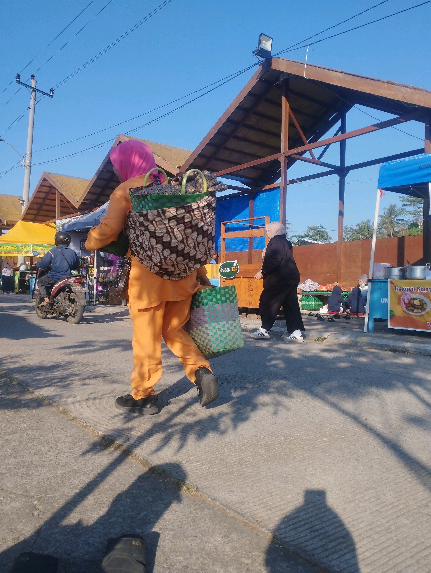The image depicts an outdoor market scene in Salatiga, Indonesia, under a clear blue sky. A person wearing an orange outfit and a pink headscarf is walking away from the camera, carrying two large bags, one on their back and another in their hand. The bag on their back is a woven basket with a brown and white patterned fabric inside. The bag in their hand is a green and white checkered basket.
In the background, a brown wooden structure with a pitched roof is visible, potentially a market stall or shelter. To the left, a yellow tent is set up, and further back, a motorcycle rider wearing a blue jacket and a black helmet is seen. To the right, another market stall with a blue tarp is present, displaying food items. Two people are seated near this stall. The ground is paved with concrete, and shadows from trees are cast across it. The overall scene suggests a bustling market environment.