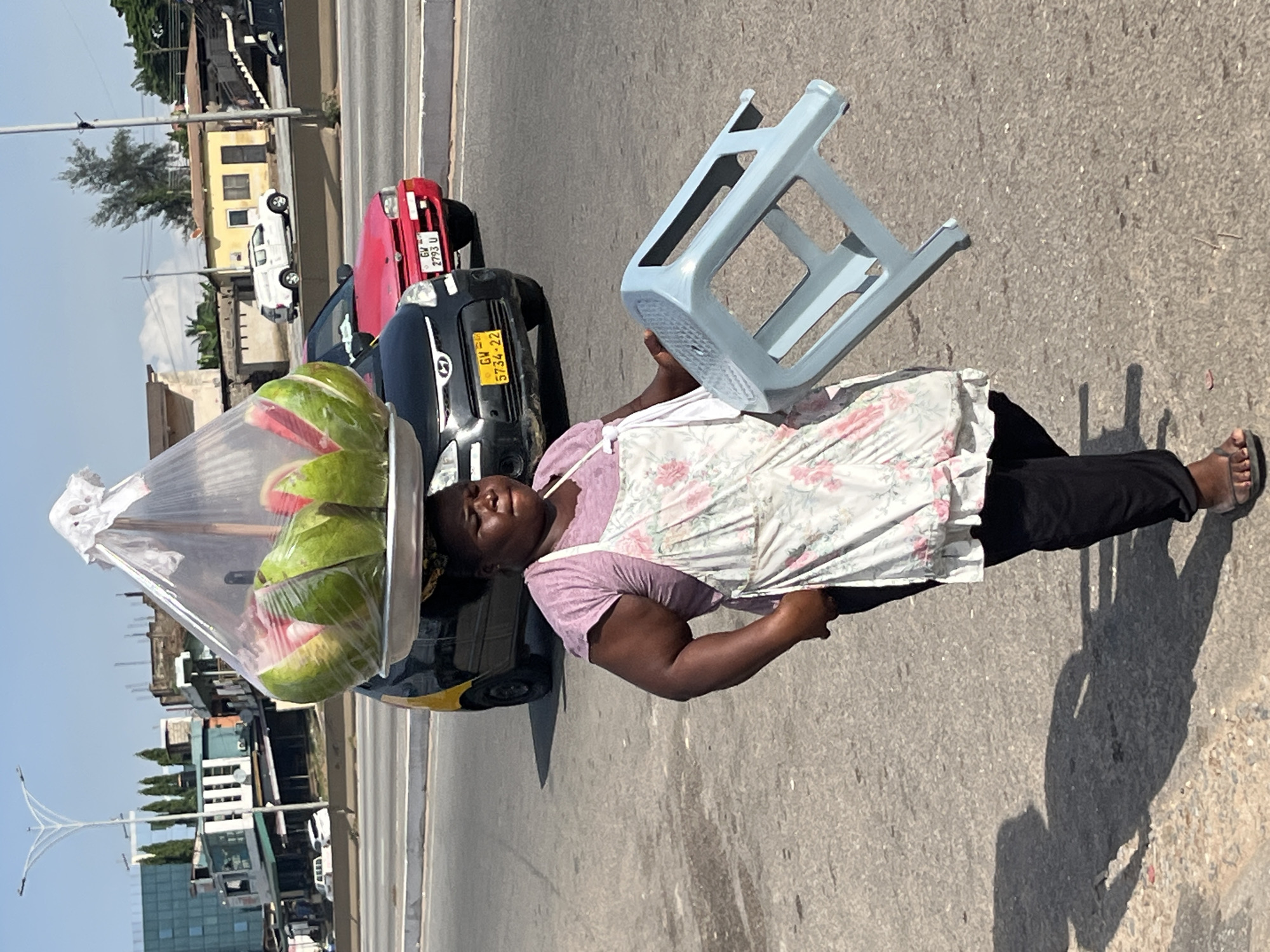 A woman, identified as a vendor, walks along a paved road in Accra, Ghana. She is carrying a tray of sliced watermelons on her head, covered in plastic wrap. In her right hand, she holds a light blue plastic stool. She is wearing a pink t-shirt, a floral apron, black pants, and flip-flops. The background shows a busy street with cars, buildings, and trees, suggesting an urban environment. The road is asphalt, and the lighting indicates it is daytime with a clear sky. The woman's shadow is cast on the ground to her left.