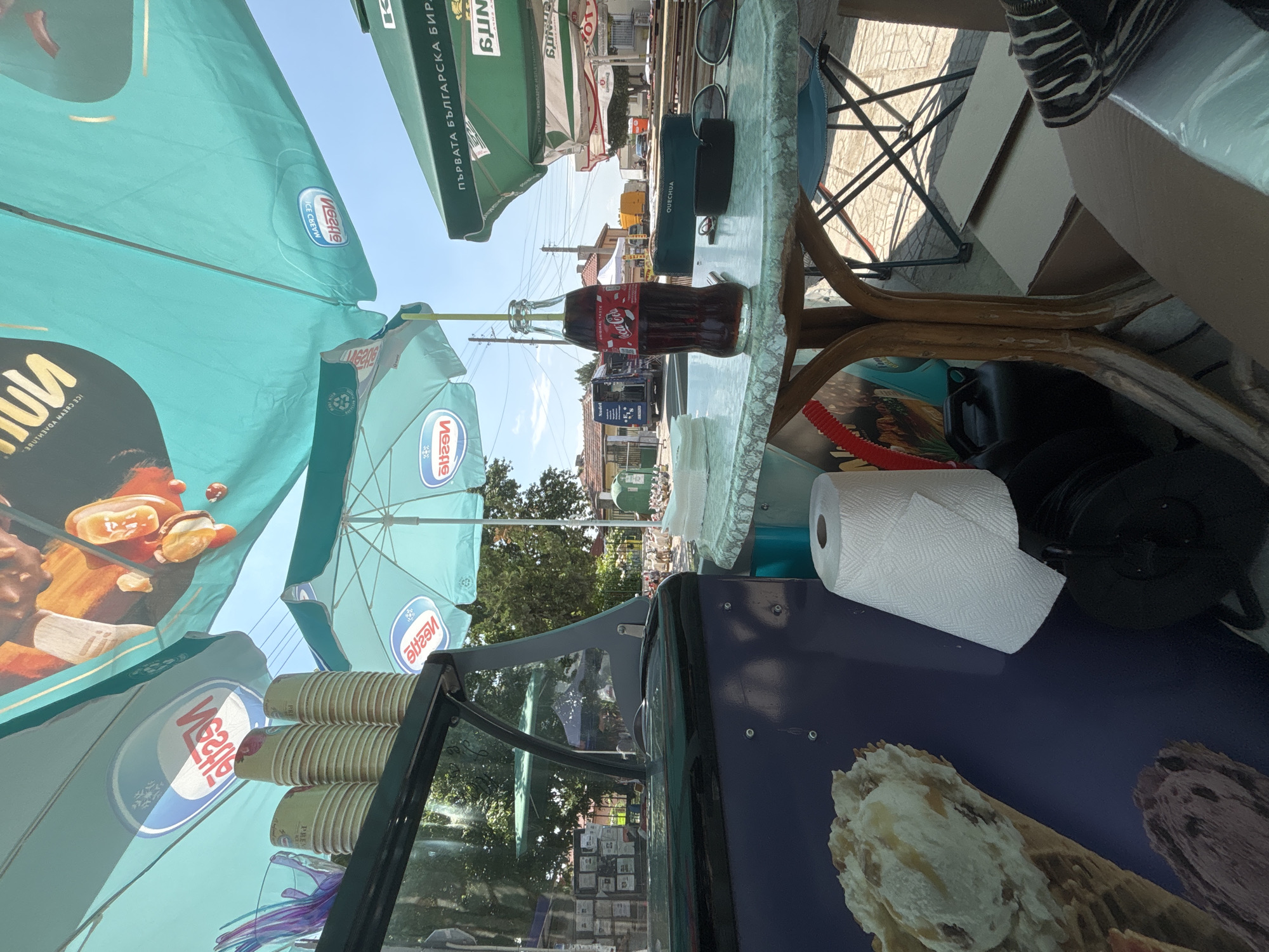The image captures an outdoor vendor scene in Kostievo, Bulgaria, under bright daylight. A light green, patterned round table with a wooden pedestal base occupies the central foreground. On this table, a glass bottle of Coca-Cola with a yellow straw stands prominently, accompanied by dark sunglasses, a black sunglasses case, and a dark green "QUECHUA" branded pouch. A roll of white paper towels is positioned to the left of the table.
To the immediate left, a portion of a dark-colored ice cream display stand is visible, featuring a slanted glass top. Stacked paper cups rest on its upper surface. The lower facade of the stand displays graphics of ice cream: a waffle cone with white and caramel-swirled ice cream, and a darker ice cream item below it.
Overhead, several large promotional umbrellas provide shade. Multiple teal-colored umbrellas feature mirrored "Nestlé" logos and "ICE CREAM" text, indicating an ice cream product. To the right, a dark green umbrella displays Bulgarian text "ПЪРВАТА БЪЛГАРСКА БИРА" (The First Bulgarian Beer) and "SUN".
The background reveals a street environment with buildings, dense foliage, and utility poles under a clear blue sky. A blue truck, branded "Nadel" with additional Bulgarian text, is parked on the left side of the street. Further down, a yellow van is visible. In the mid-ground right, a group of white fowl, possibly geese or chickens, congregates near a low green structure. No individuals are present in the visible frame.