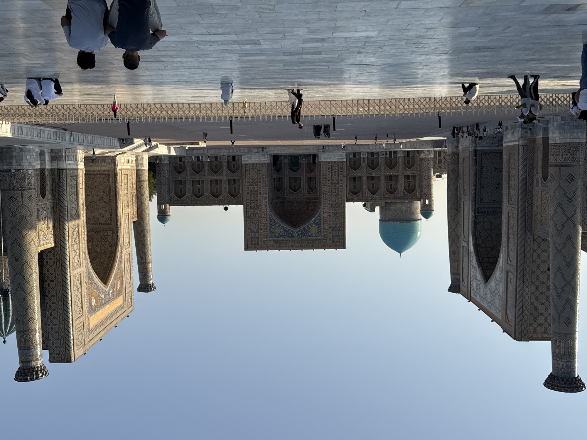 This is a full, eye-level, wide shot of the Registan Square in Samarkand, Uzbekistan, during daylight with a clear, pale blue sky. The image captures the grand scale of the three madrasahs that form the square: the Ulugh Beg Madrasah, the Sher-Dor Madrasah, and the Tilla-Kari Madrasah. The central and furthest madrasah is Ulugh Beg, with its iconic turquoise dome. To its left is the Sher-Dor Madrasah, and to its right is the Tilla-Kari Madrasah. Both Sher-Dor and Tilla-Kari have towering minarets on either side of their main entrances. The facades of the madrasahs are richly decorated with intricate geometric patterns and calligraphy in blue, gold, and white tiles.
In the foreground, the paved courtyard of the Registan is visible, with a section of light-colored, large, square tiles. Several people are scattered across the courtyard. In the immediate foreground, two men are seated with their backs to the camera, looking towards the madrasahs. To their right, a group of three people, two men and one woman, are seated, with two of them looking towards the camera. Further back, a young child in a pink shirt is walking away from the camera. A woman wearing a light blue headscarf is seated to the right of the child. A group of four people, two men and two women, are walking away from the camera on the right side of the frame.
In the middle ground, there is a low, ornate fence or railing separating the foreground seating area from the main plaza. Beyond the fence, a number of tourists and locals are walking and standing. Some are observing the architecture, while others are engaged in conversation. A few figures are seen in the arched entrances of the madrasahs. The overall atmosphere is one of calm tourism and appreciation of historical architecture. The sunlight casts some shadows, indicating it is likely late afternoon.