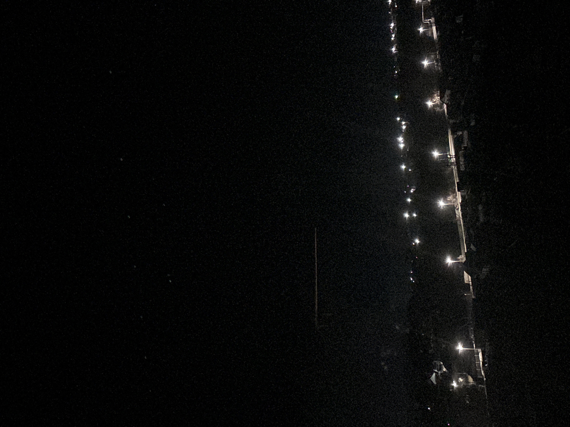The image shows a dark, nighttime scene in what appears to be an urban or suburban area of Chaikovsky, Russia. Along the right side of the image, a series of illuminated streetlights are visible, suggesting a road or walkway. The left side of the image is dominated by darkness, with little to no discernible detail except for some faint objects in the middle area. The image is grainy, potentially due to low-light conditions.