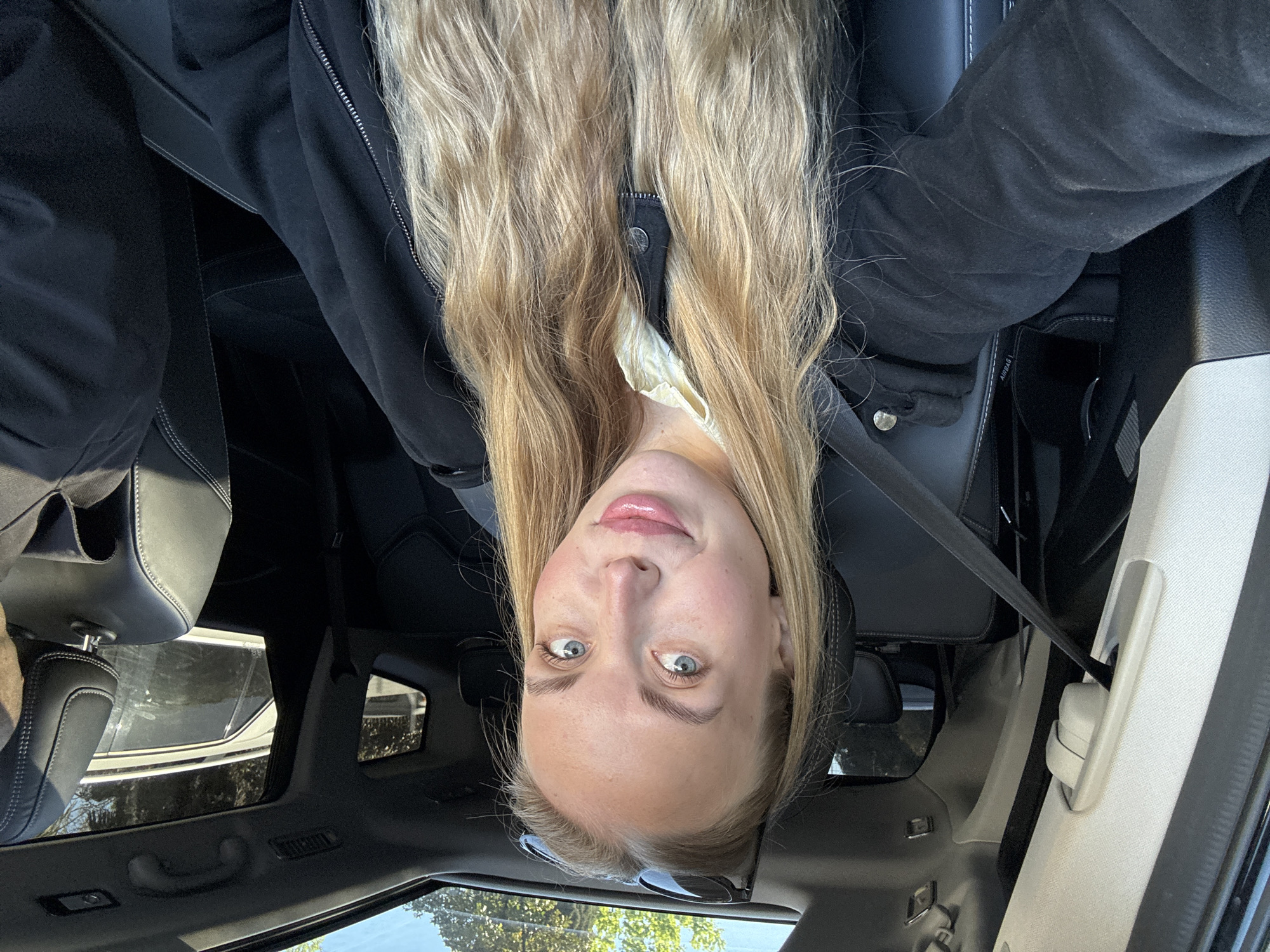 A portrait-oriented selfie captures a young woman with long, wavy blonde hair and blue eyes, smiling subtly as she looks directly at the camera. The original image is inverted, showing her upside down. She is seated inside a car, wearing a black zippered jacket over a light-colored top, with a seatbelt visible across her chest.
The interior of the car features black leather seats with white stitching, and a light-colored ceiling. A sunroof or panoramic roof is visible above, allowing natural light to filter in and offering a glimpse of trees and a bright sky outside, indicating it is daytime. On the side of a seat, the word "AIRBAG" is visible. The scene suggests a casual moment, possibly taken while on a journey or parked in Sochi, Russia.