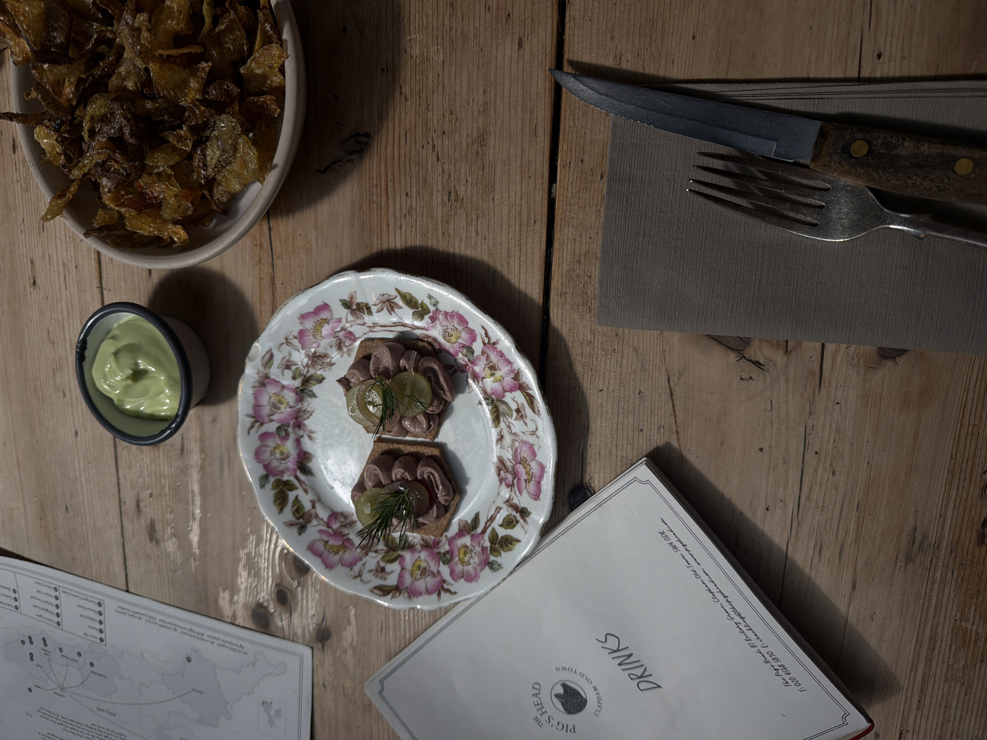 The image depicts a wooden table setting with a variety of items related to dining. At the top left, there is a bowl filled with what appears to be crispy potato chips. Adjacent to it, there is a small cup of green sauce. In the center of the image, there is a decorative floral plate with two servings of what appears to be pate on toast, garnished with greens. To the right of the plate, there is a fork and knife placed on a neutral-toned napkin. Below the plate, there is a "DRINKS" menu from "THE PIG'S HEAD CLAPHAM OLD TOWN". Adjacent to the menu, there is a map of England printed on paper. The text on the menu indicates the location as 87 Rectory Grove, Clapham Old Town, SW4 ODR, which is located in London, United Kingdom.